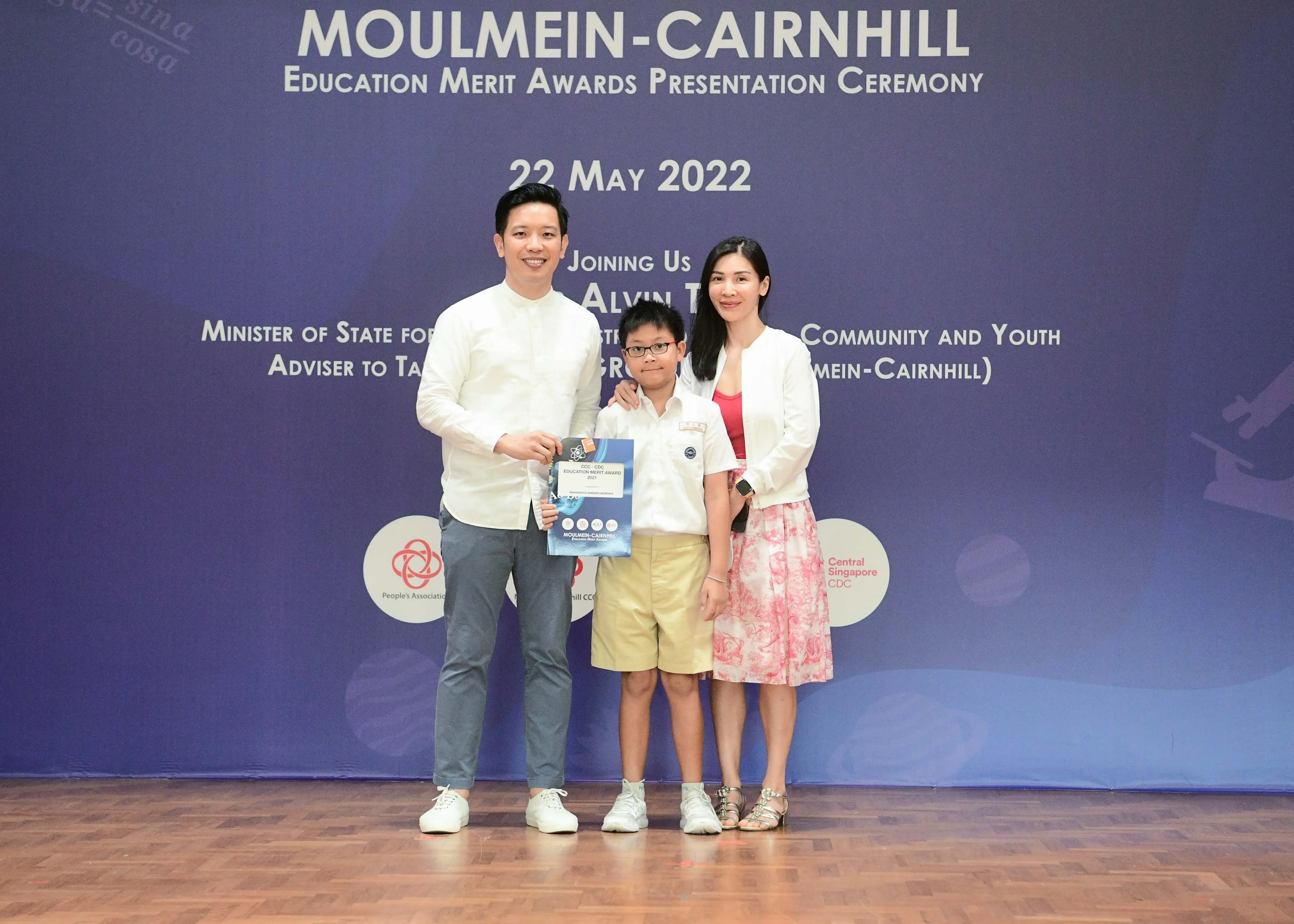 Two adults and a child holding an award certificate at the Moulmein-Cairnhill Education Merit Awards ceremony on 22 May 2022.