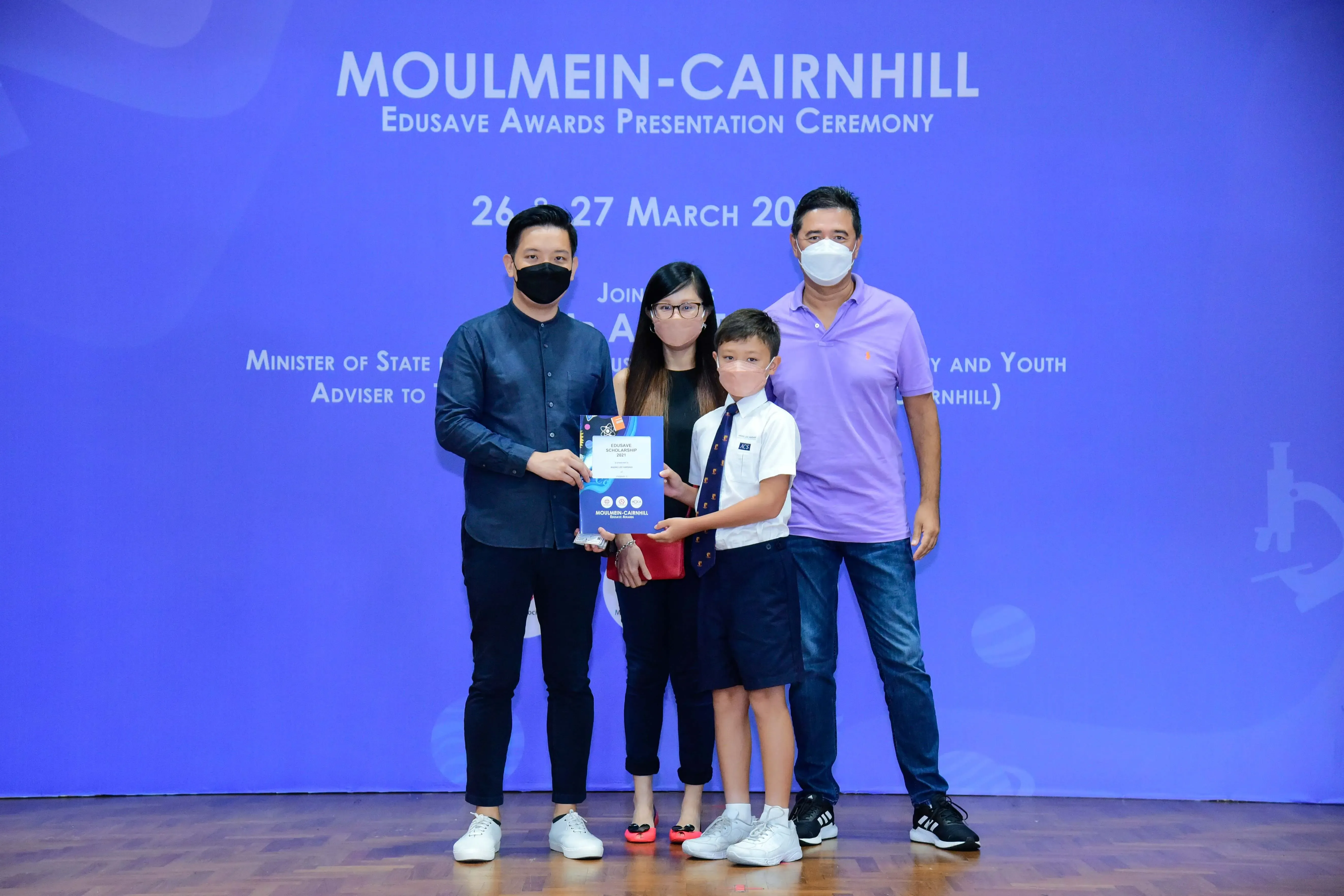 Four masked people pose at the Moulmein-Cairnhill Edusave Awards Presentation Ceremony, holding a certificate.