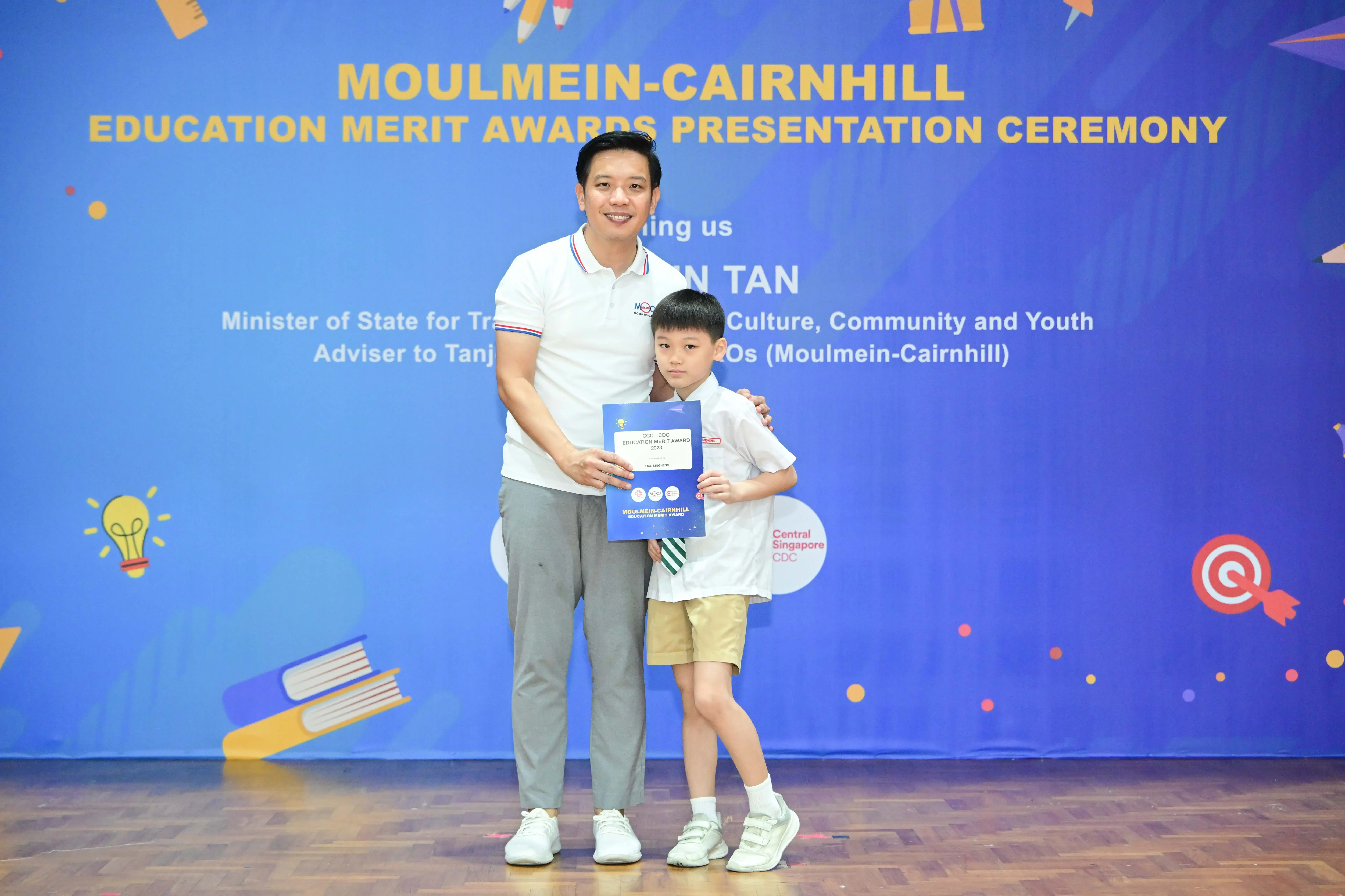 A man and young boy pose holding a certificate in front of a Moulmein-Cairnhill Education Merit Awards banner.