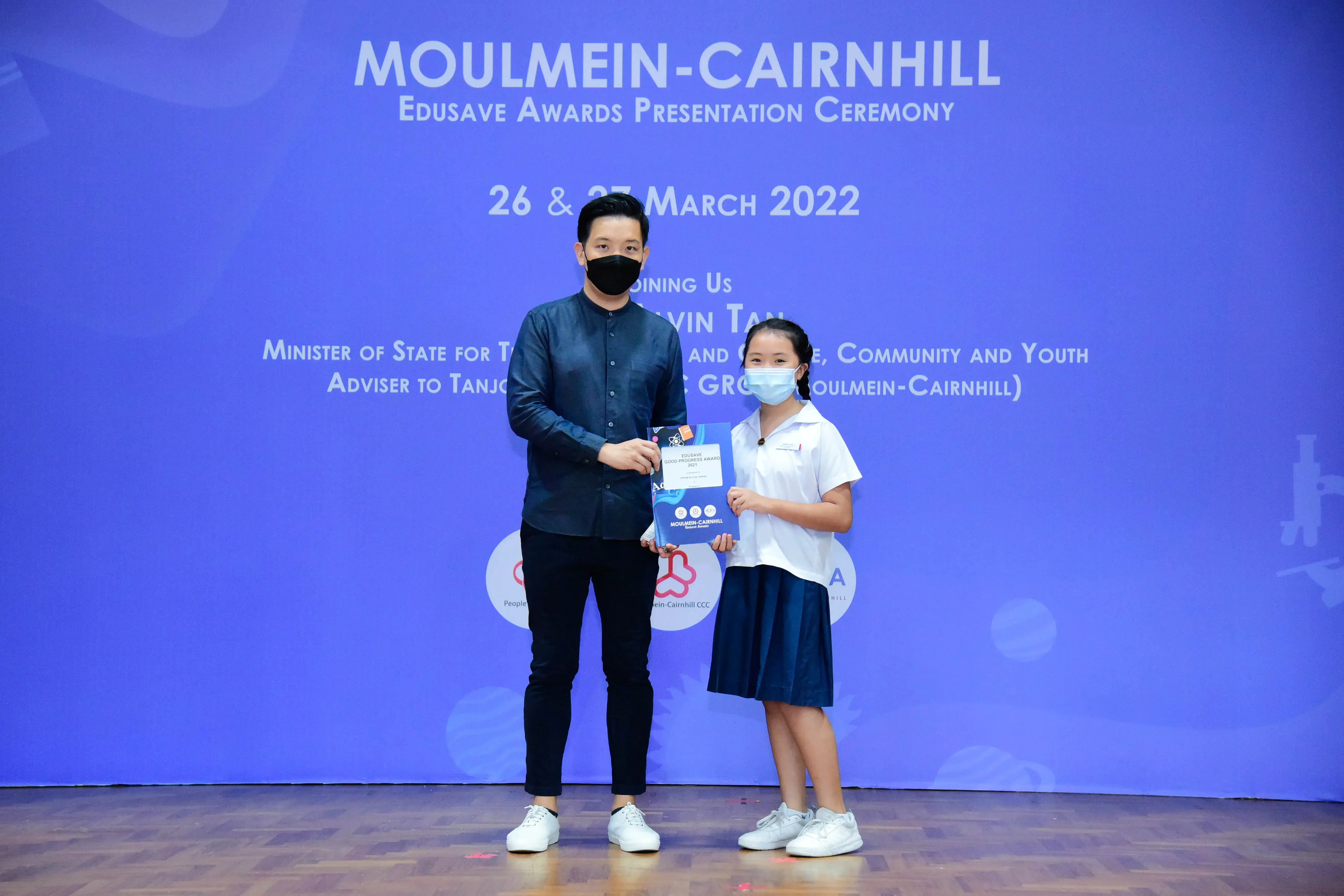 Adult and young girl in masks hold an award certificate at the Moulmein-Cairnhill Edusave Awards Ceremony 2022.