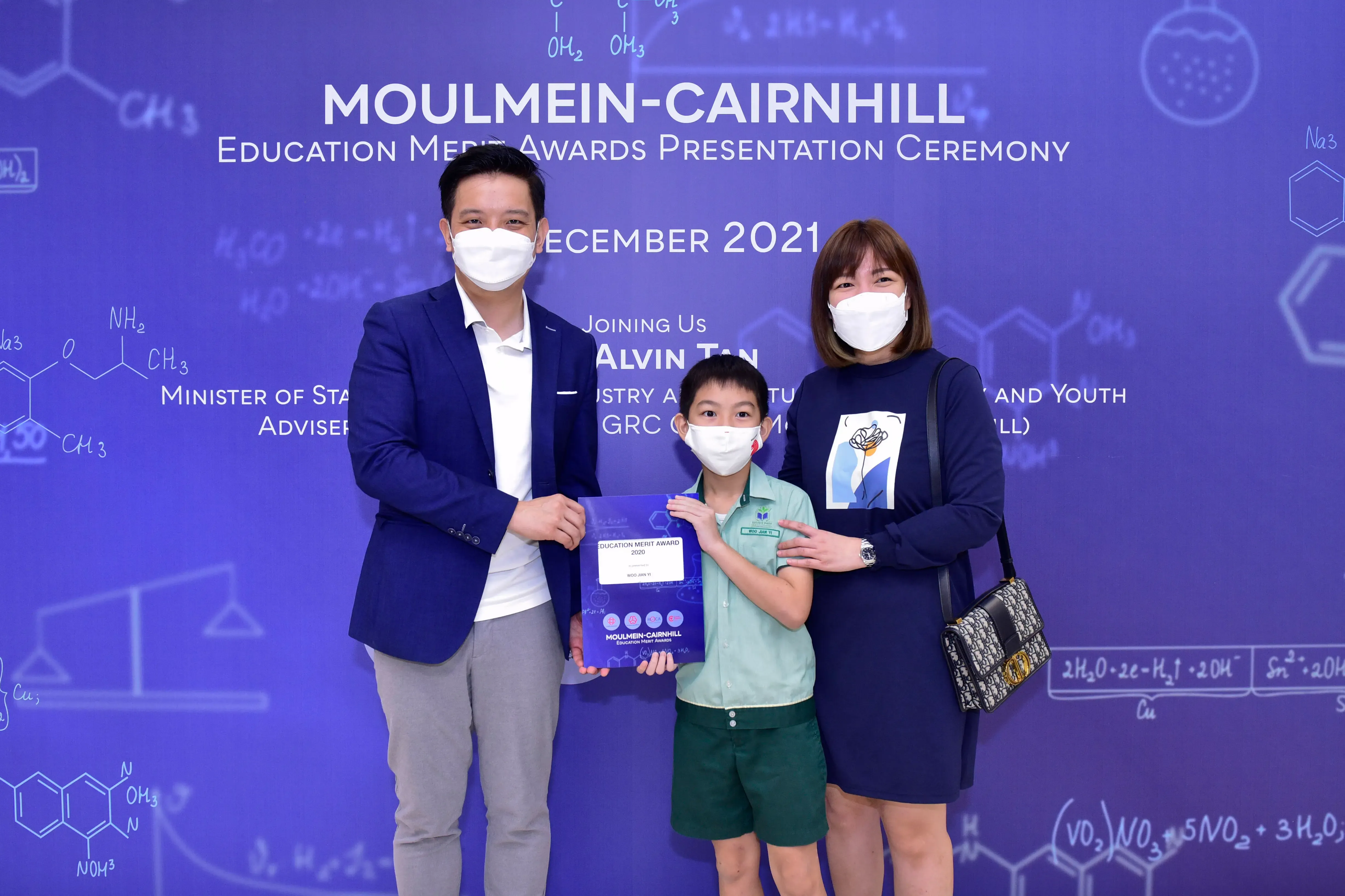 Two masked adults and a masked child holding an award certificate at the Moulmein-Cairnhill Education Merit Awards 2021.
