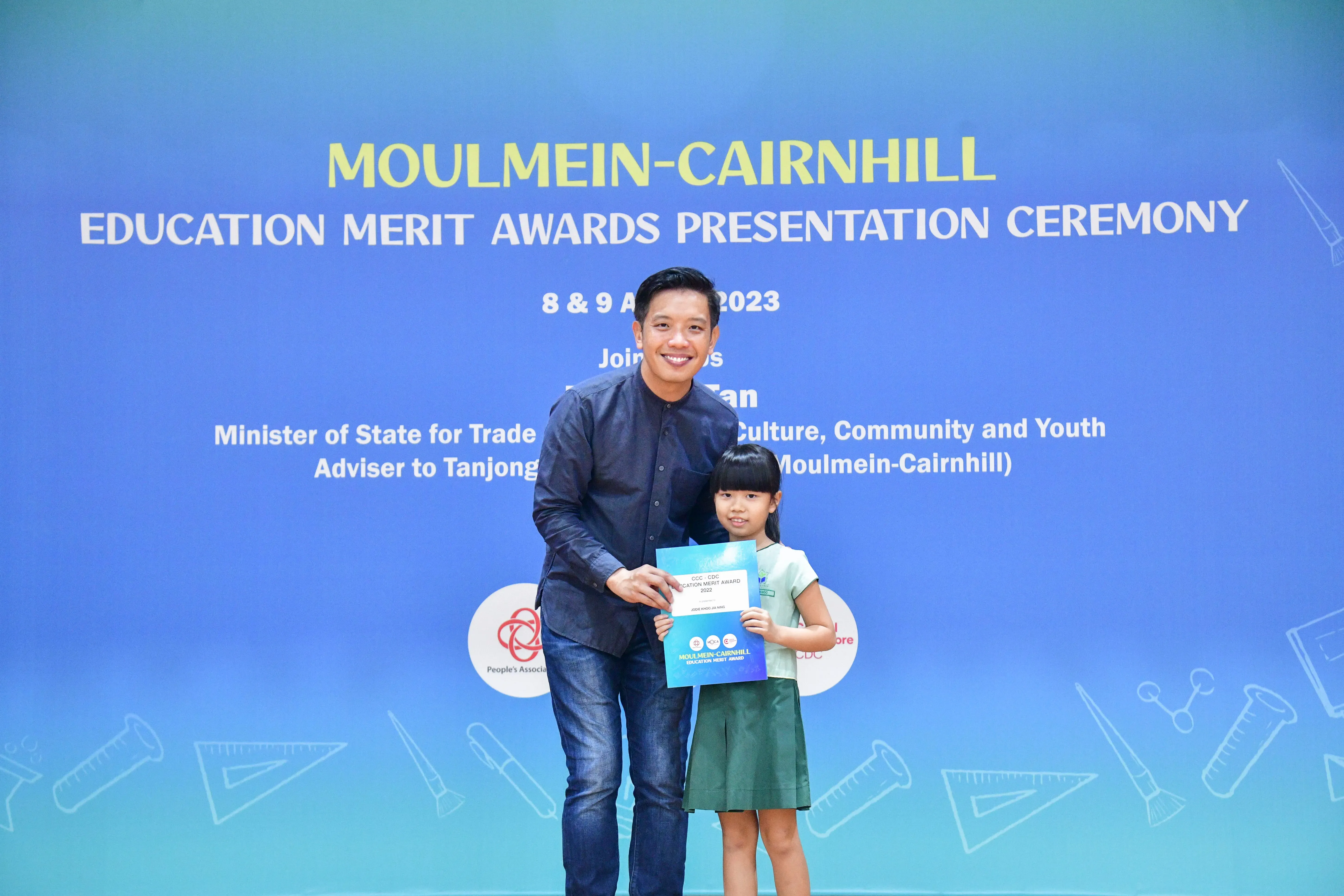 A man and a young girl hold a certificate in front of a Moulmein-Cairnhill Education Merit Awards 2023 banner.