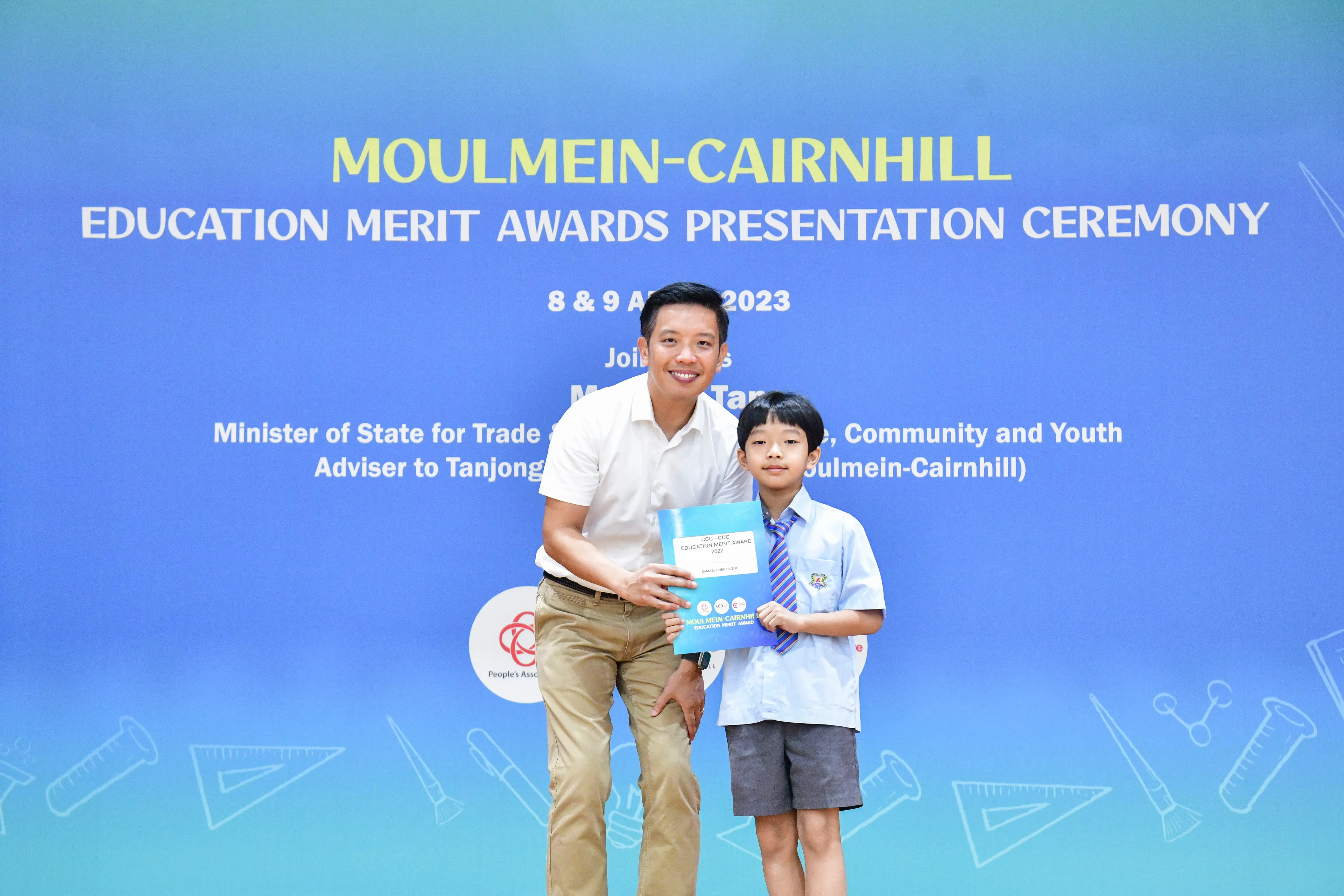 A man and a young boy in school uniform hold an award certificate in front of a Moulmein-Cairnhill Education Merit Awards banner.