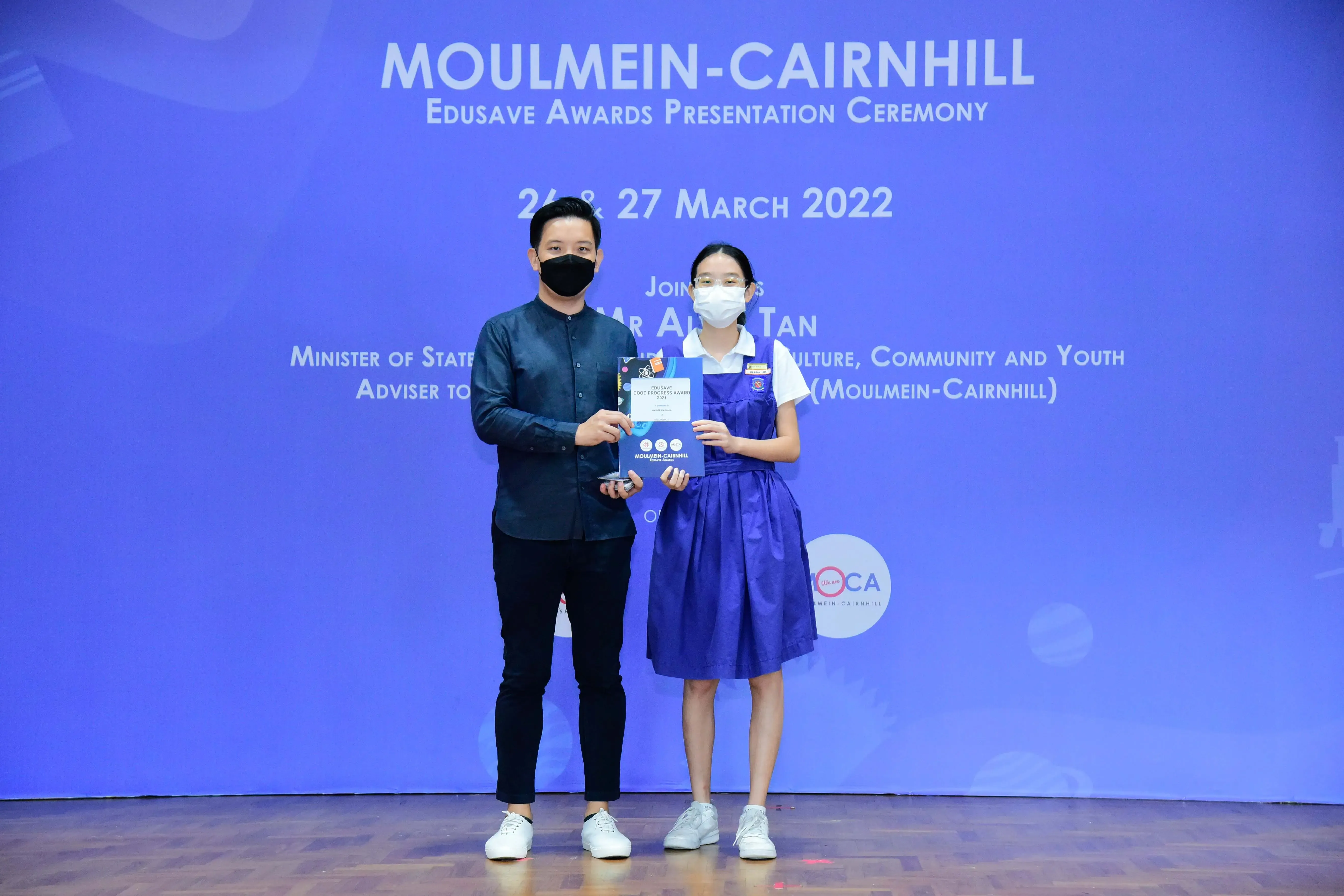 Two masked individuals hold an award certificate on stage at the Moulmein-Cairnhill Edusave Awards Ceremony, March 2022.
