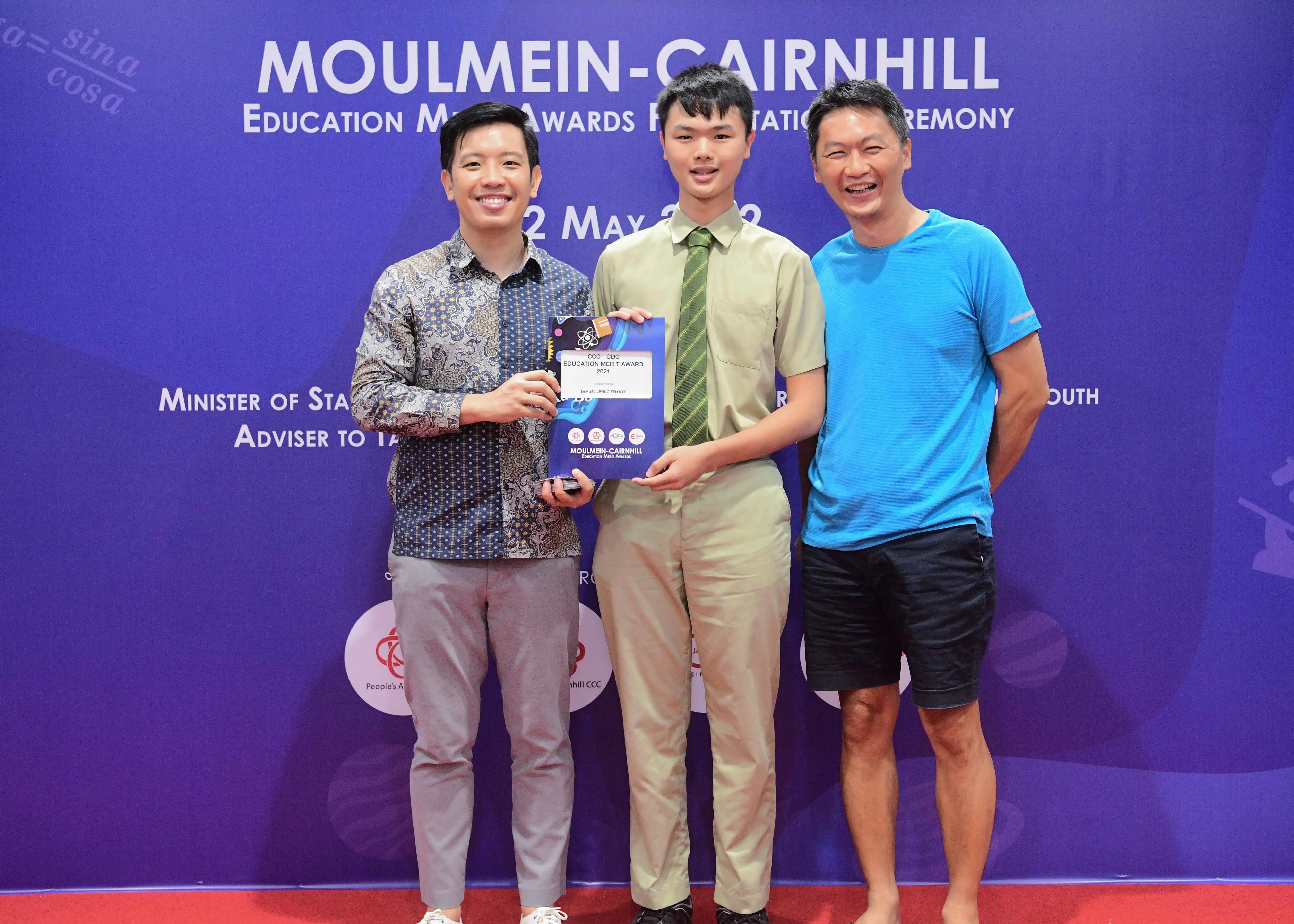 Three men smile at the Moulmein-Cairnhill Education Merit Awards ceremony, with the center man holding an award certificate.