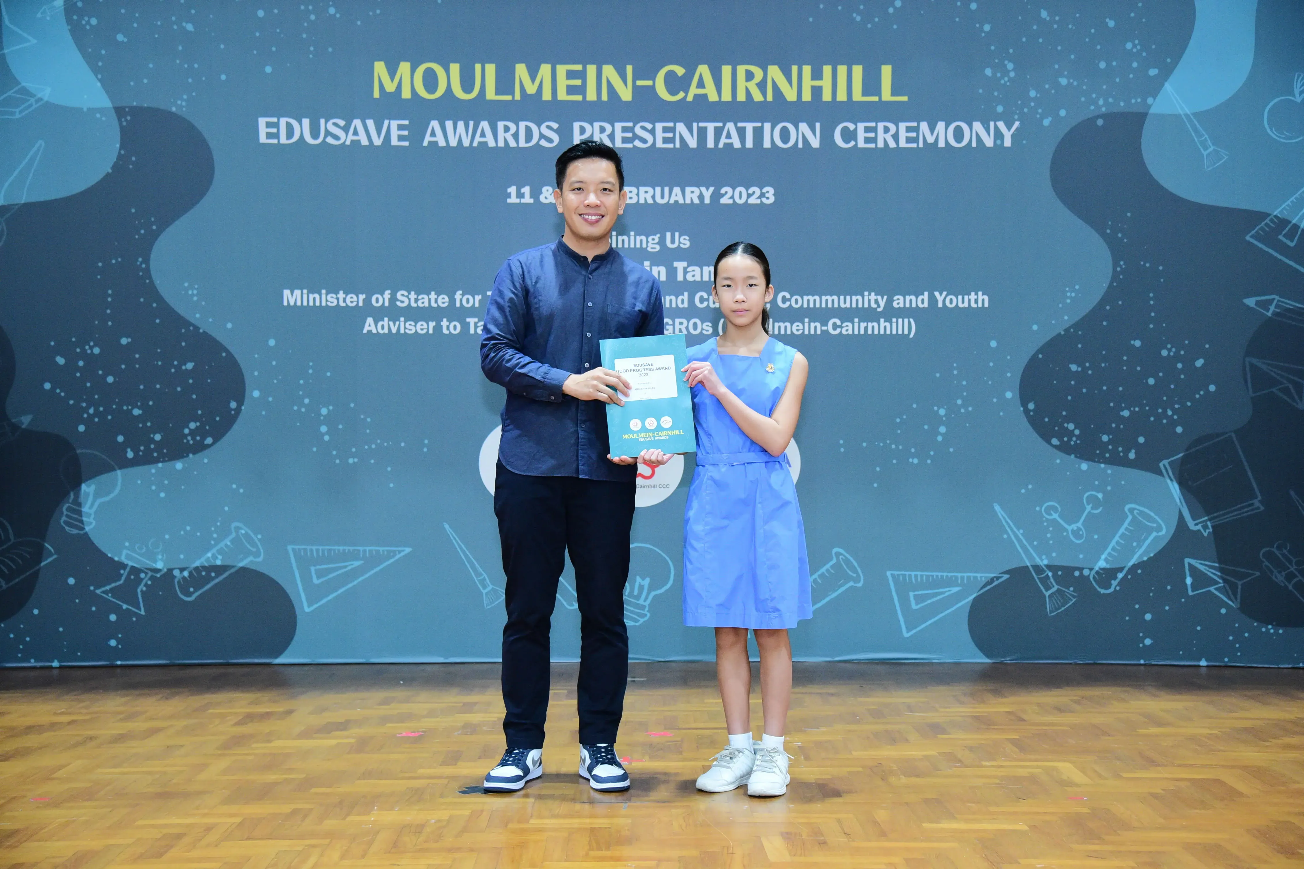 A man and a young girl hold a certificate on stage at the Moulmein-Cairnhill Edusave Awards Ceremony 2023.