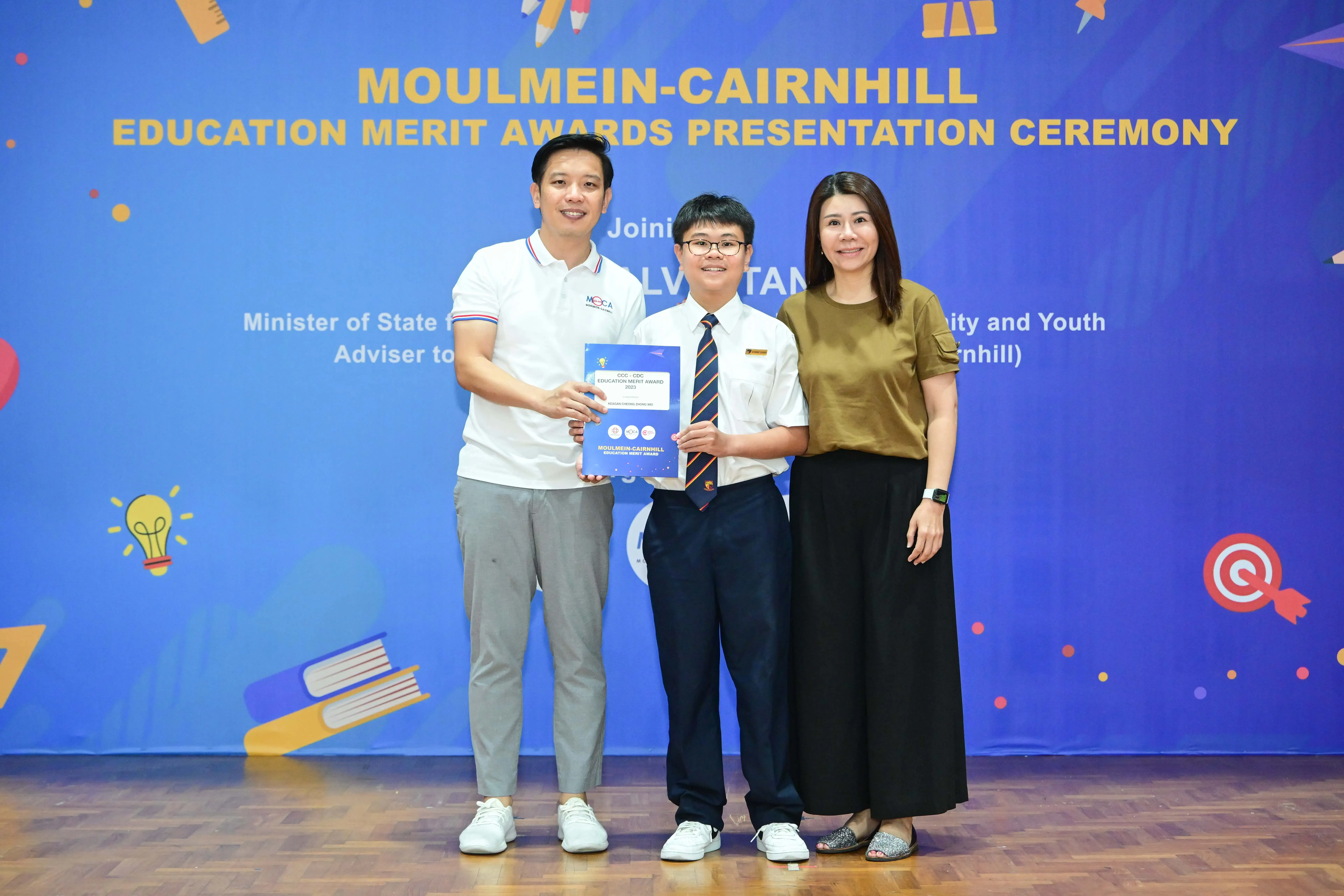 Three people pose at the Moulmein-Cairnhill Education Merit Awards Presentation Ceremony, with a student holding a certificate.