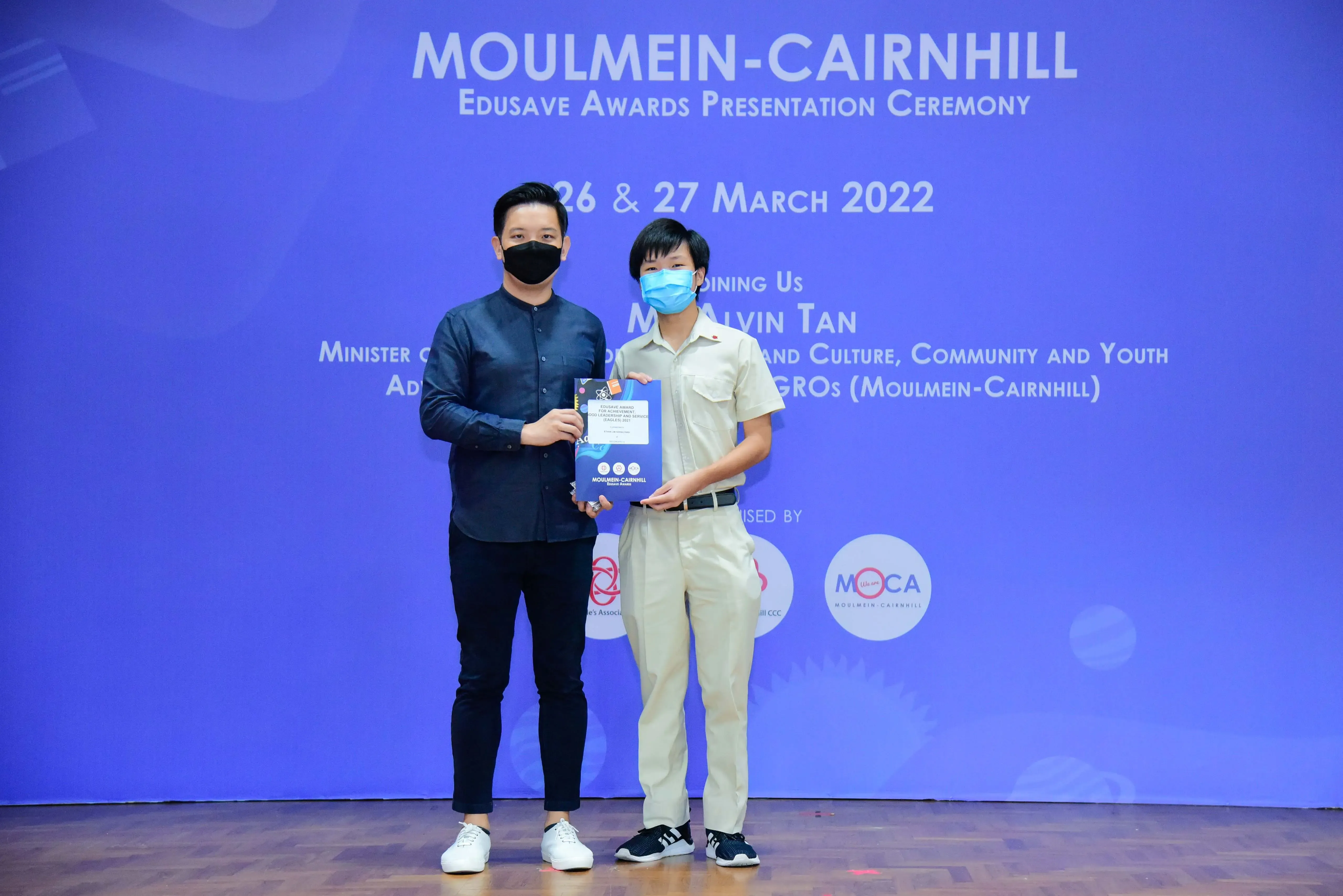 Two masked individuals hold an award at the Moulmein-Cairnhill Edusave Awards Presentation Ceremony, March 2022.