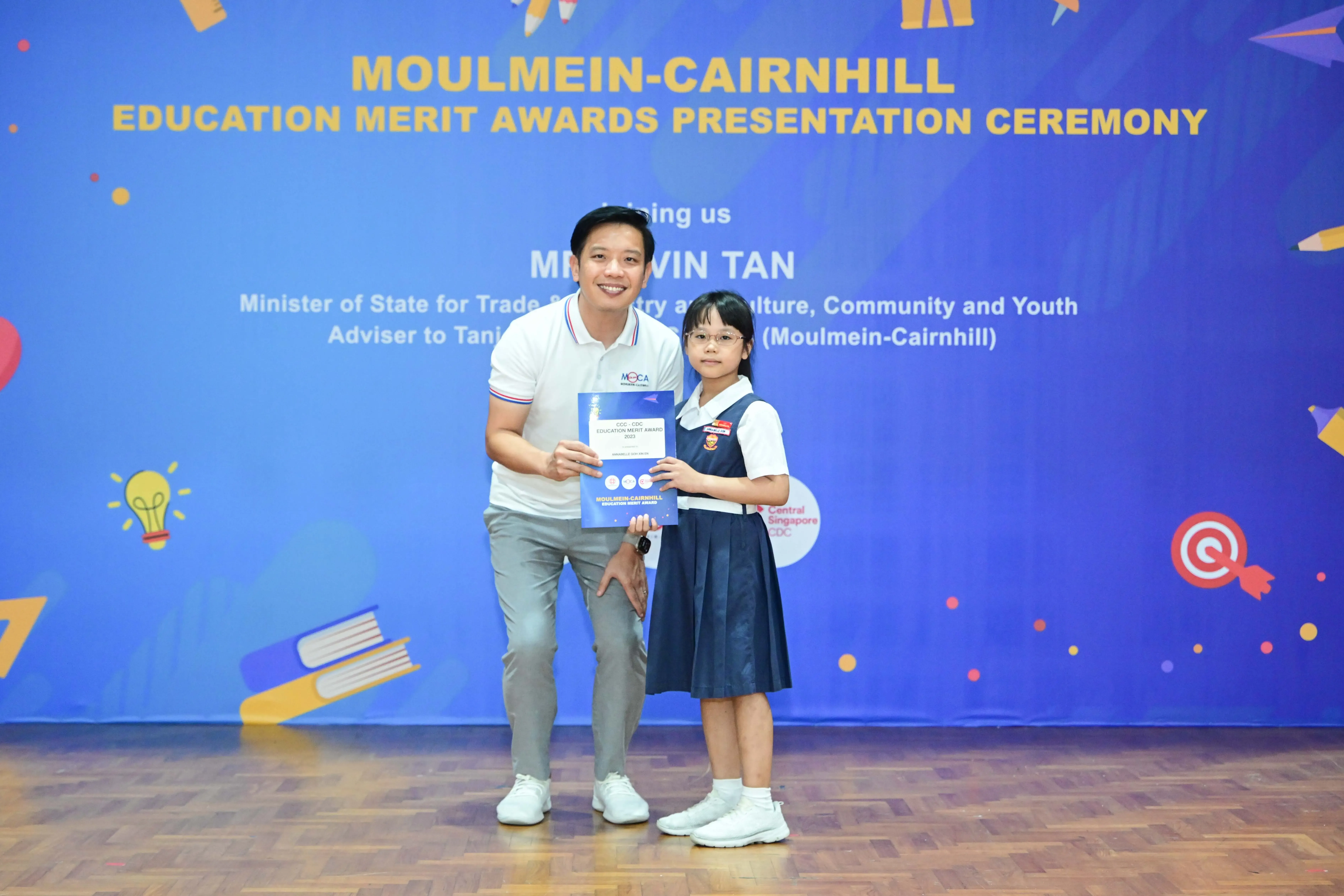 A man and a young girl in school uniform hold an award certificate at the Moulmein-Cairnhill Education Merit Awards ceremony.
