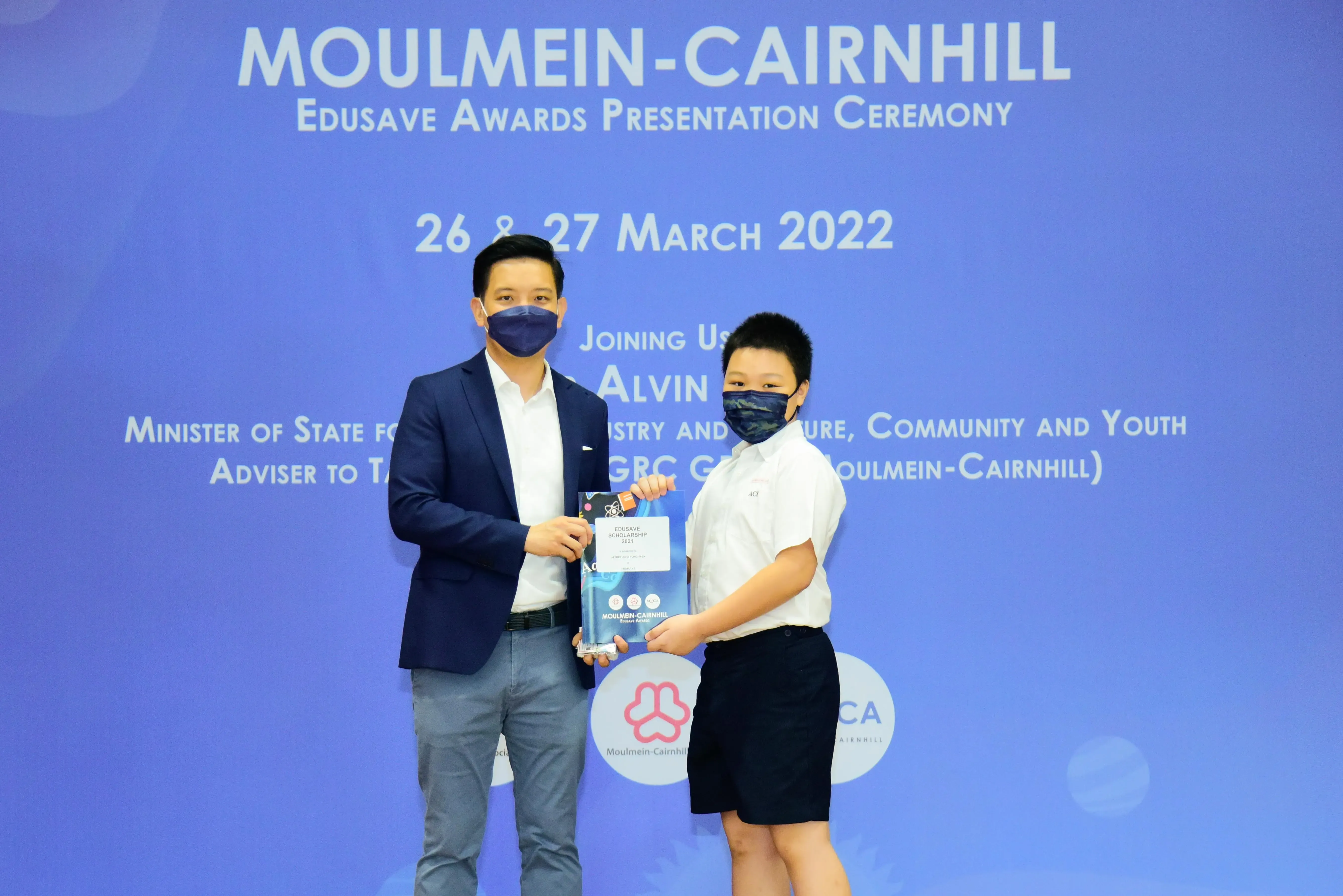 Two masked individuals hold an Edusave Scholarship award at the Moulmein-Cairnhill Awards Ceremony, March 2022.
