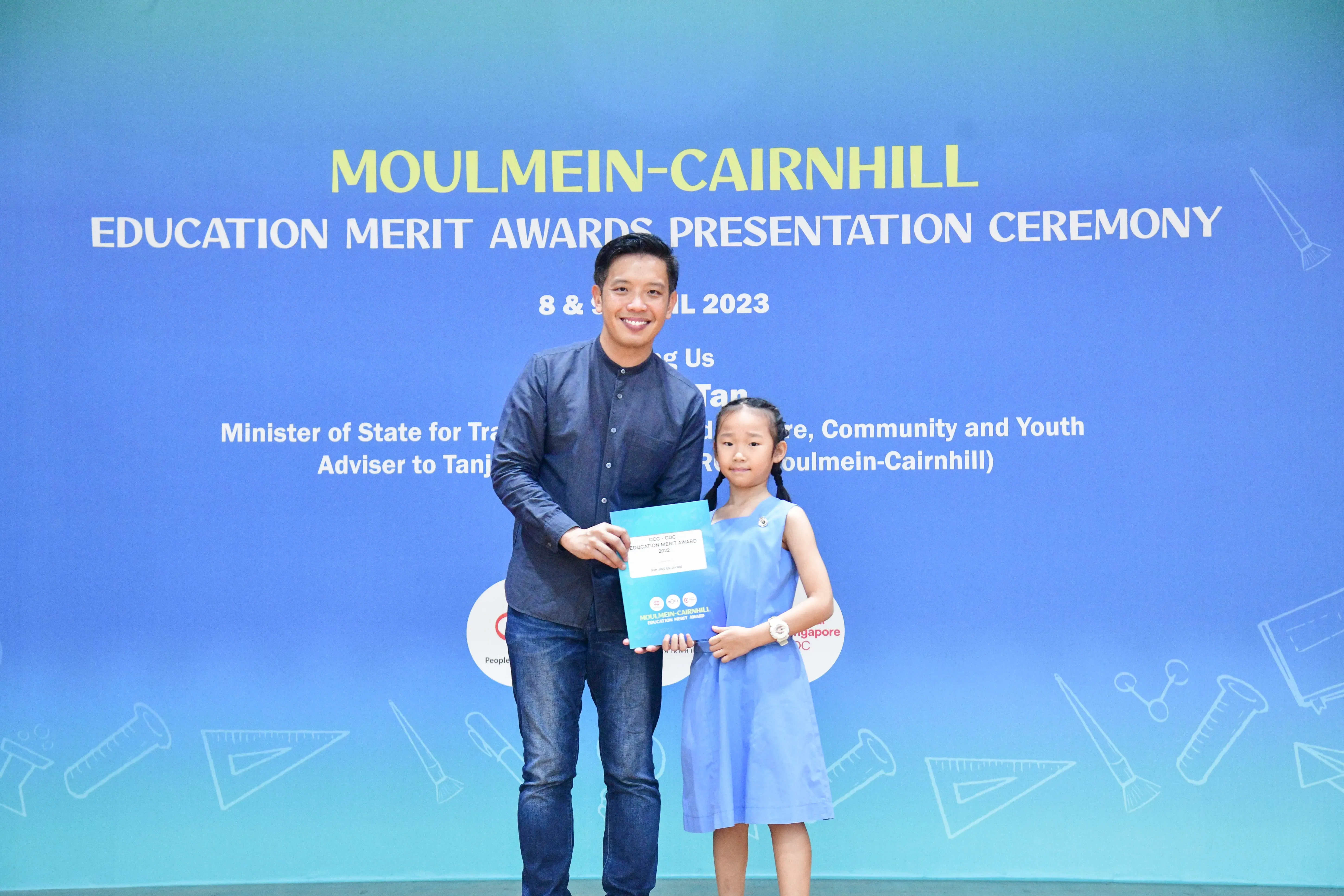 A man and a young girl hold an award certificate in front of a Moulmein-Cairnhill Education Merit Awards banner.