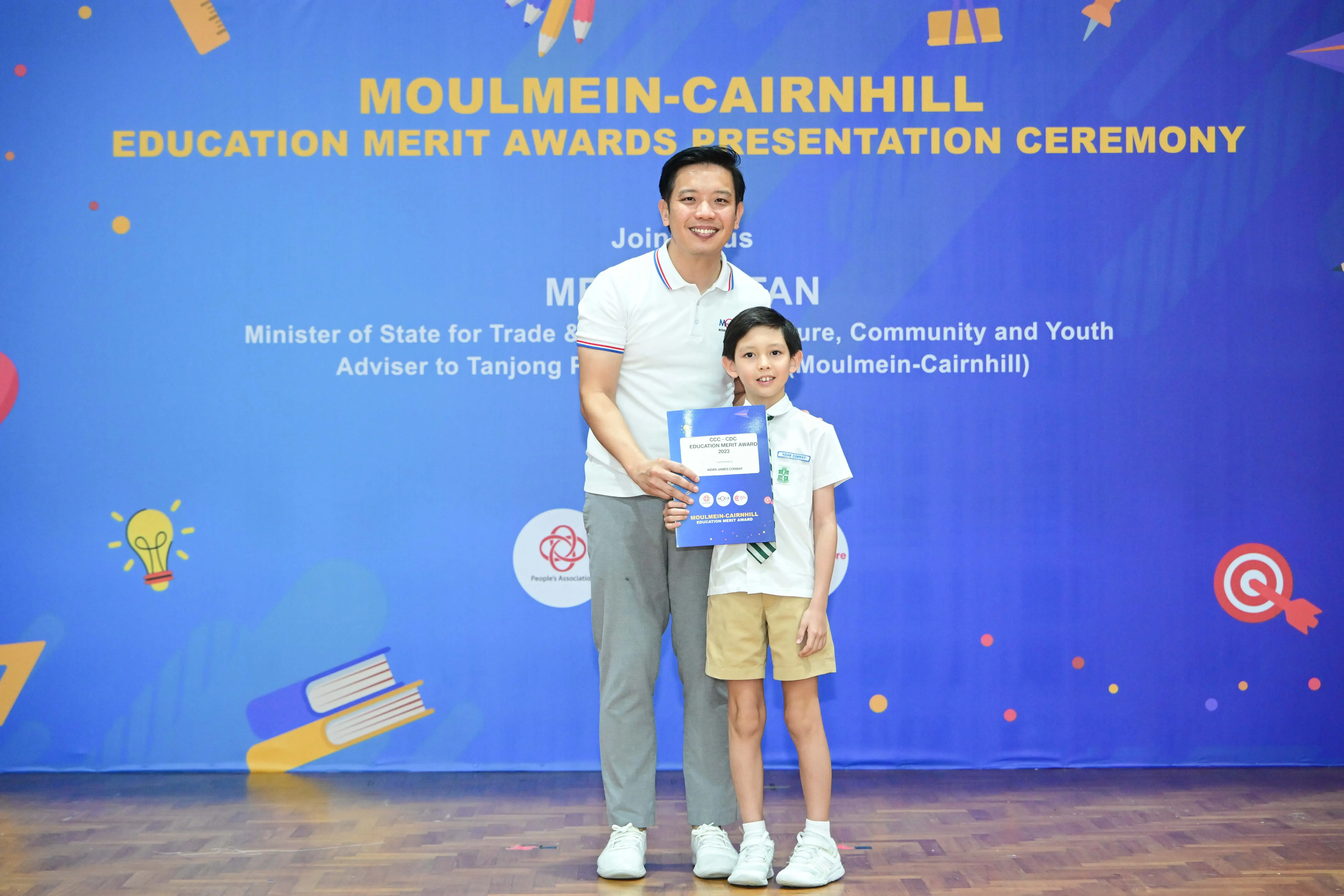 A man and a young child hold a certificate in front of a Moulmein-Cairnhill Education Merit Awards banner.