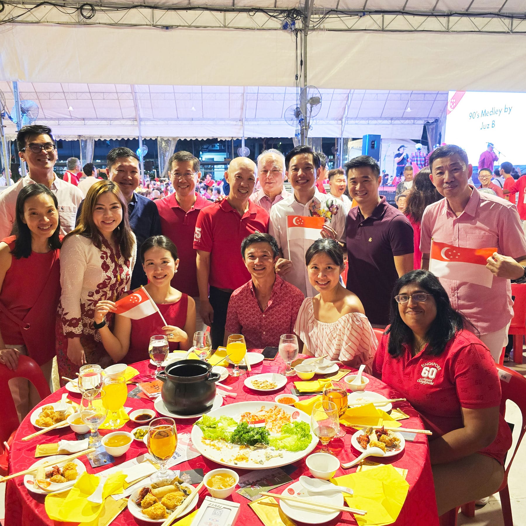 A group of smiling people in red outfits hold Singapore flags around a round table with food at an outdoor celebration.