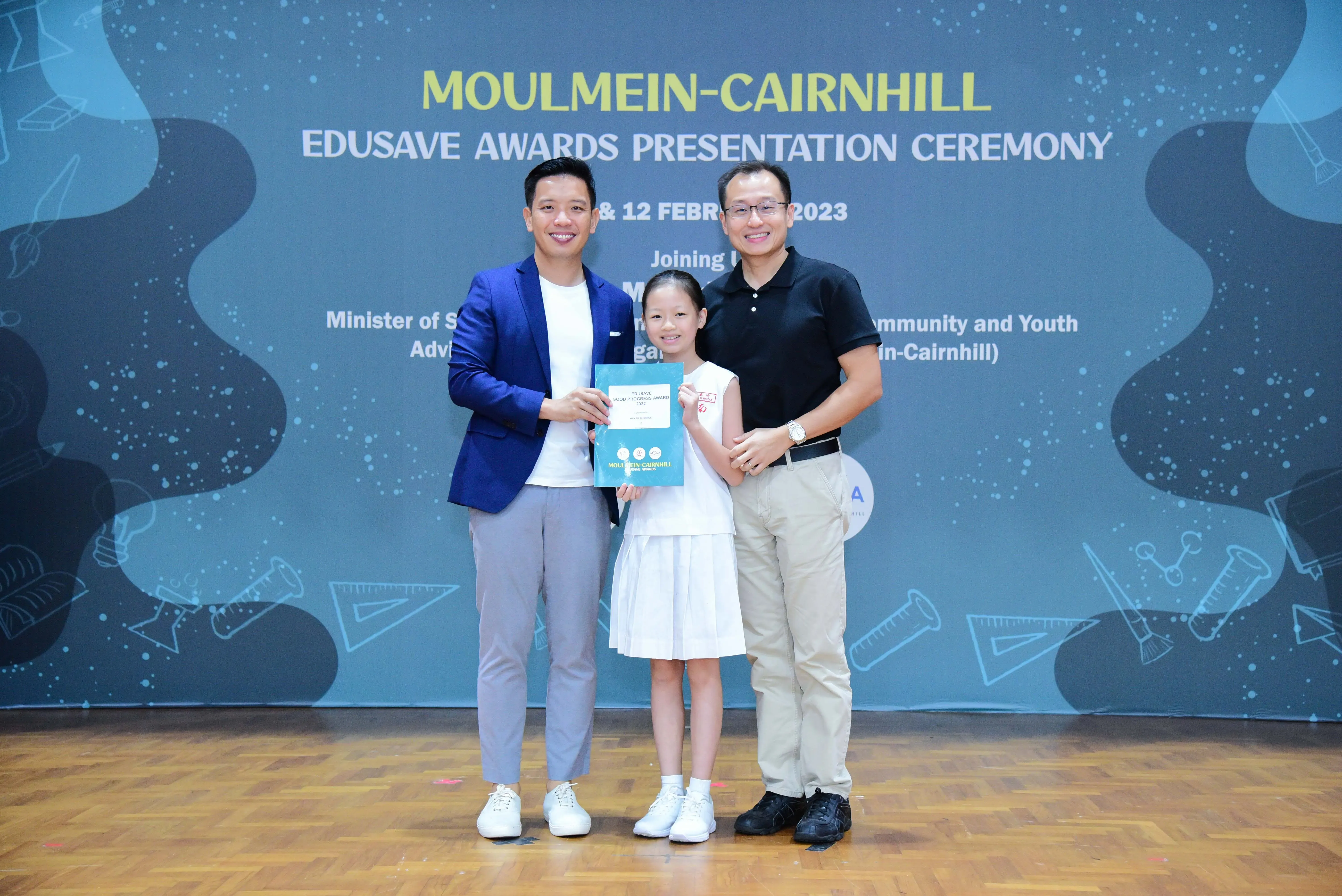 Two men and a young girl holding a certificate pose at the Moulmein-Cairnhill Edusave Awards Ceremony 2023.
