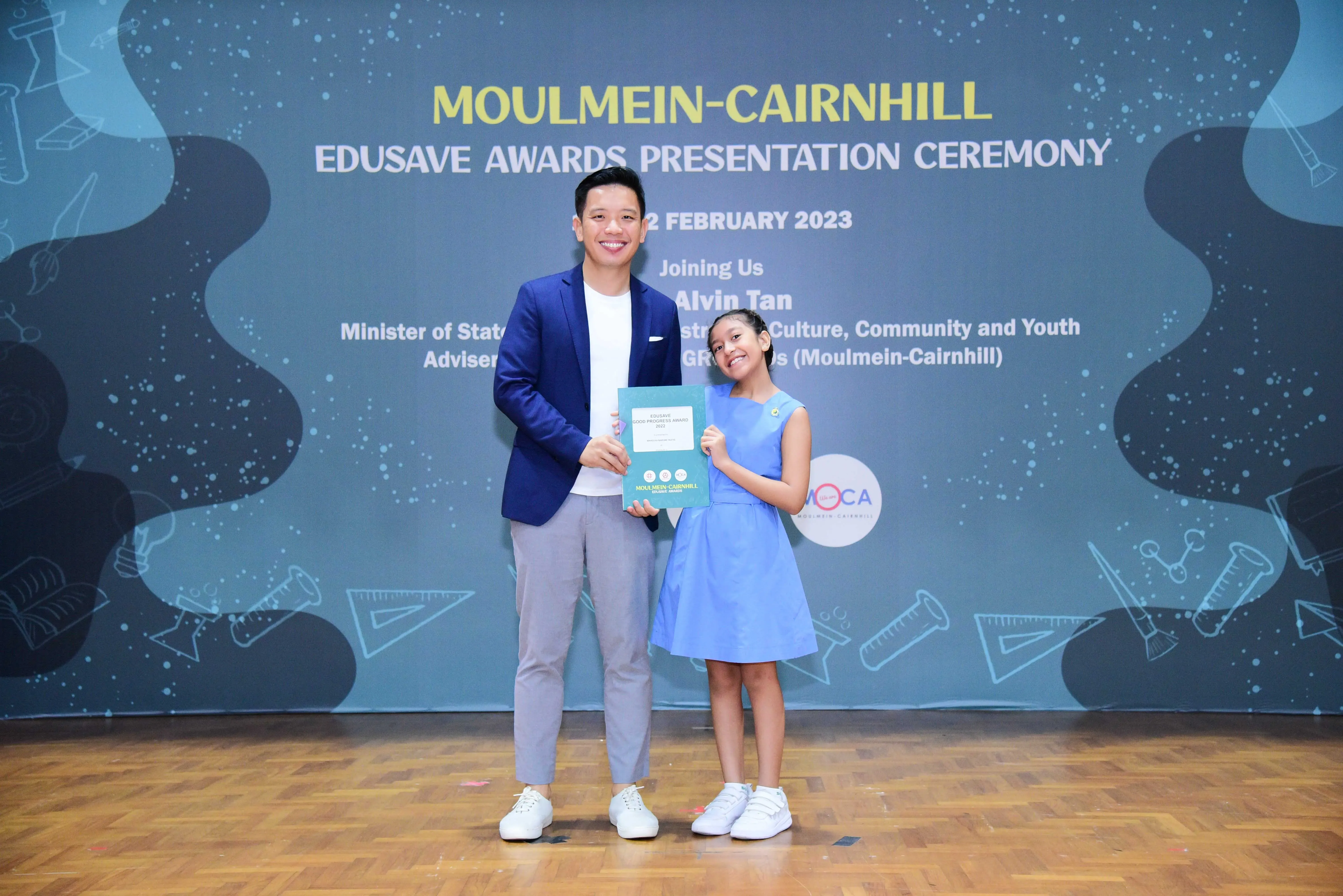 A man in a blue blazer and a young girl in a blue dress hold a certificate at the Moulmein-Cairnhill Edusave Awards 2023.
