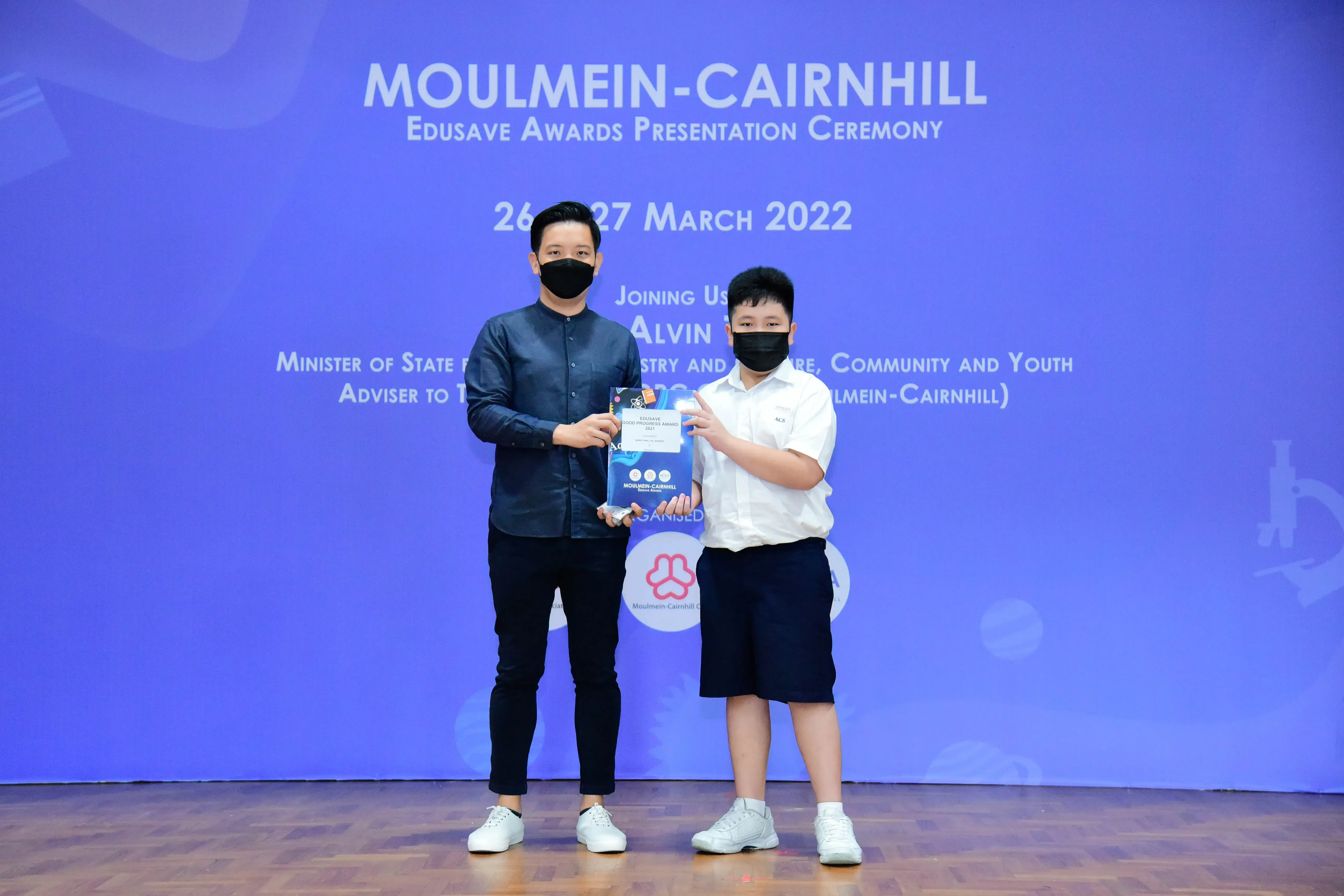 Two masked individuals hold an award booklet on stage at the Moulmein-Cairnhill Edusave Awards Ceremony, March 2022.