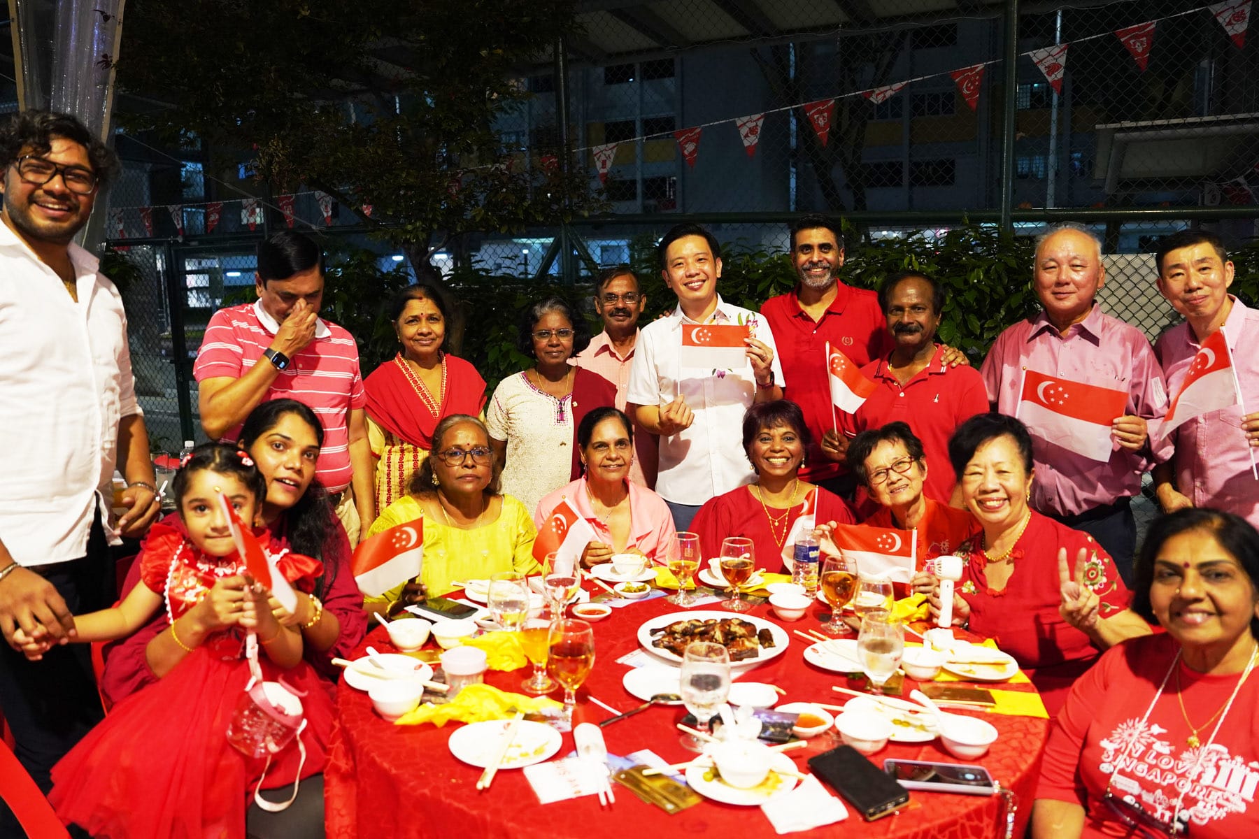 A diverse group of smiling people in red clothing hold Singapore flags at an outdoor dinner table with food and drinks.