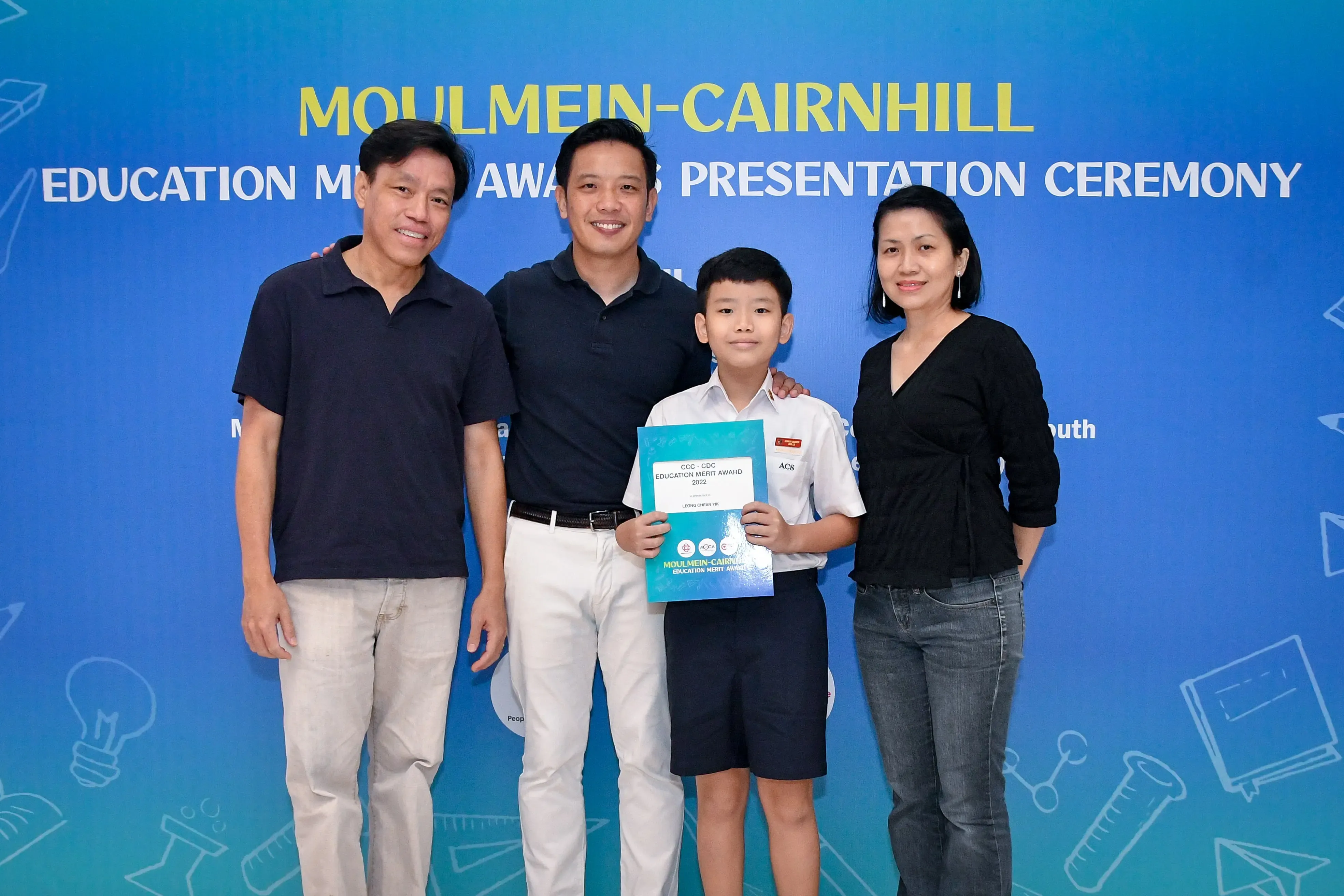 Four people pose before a Moulmein-Cairnhill Education Merit Awards banner; a boy holds a certificate.