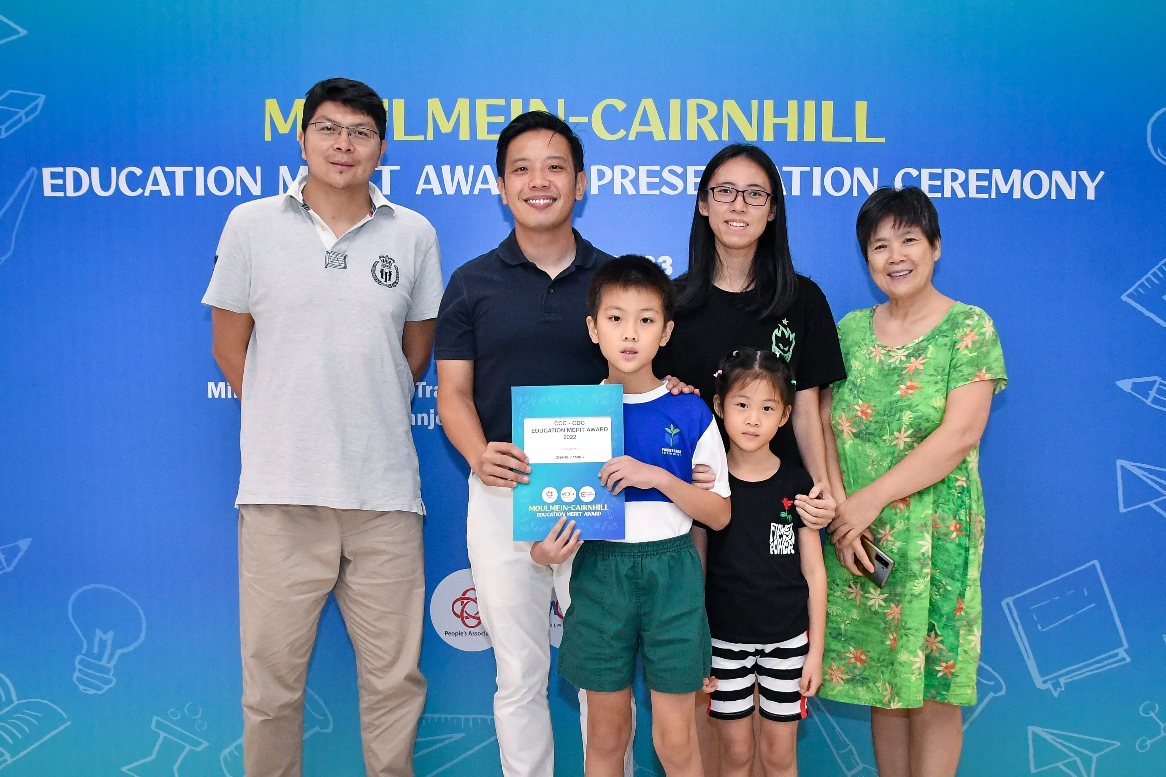 Five adults and two children pose before a Moulmein-Cairnhill Education Merit Award Presentation Ceremony banner.