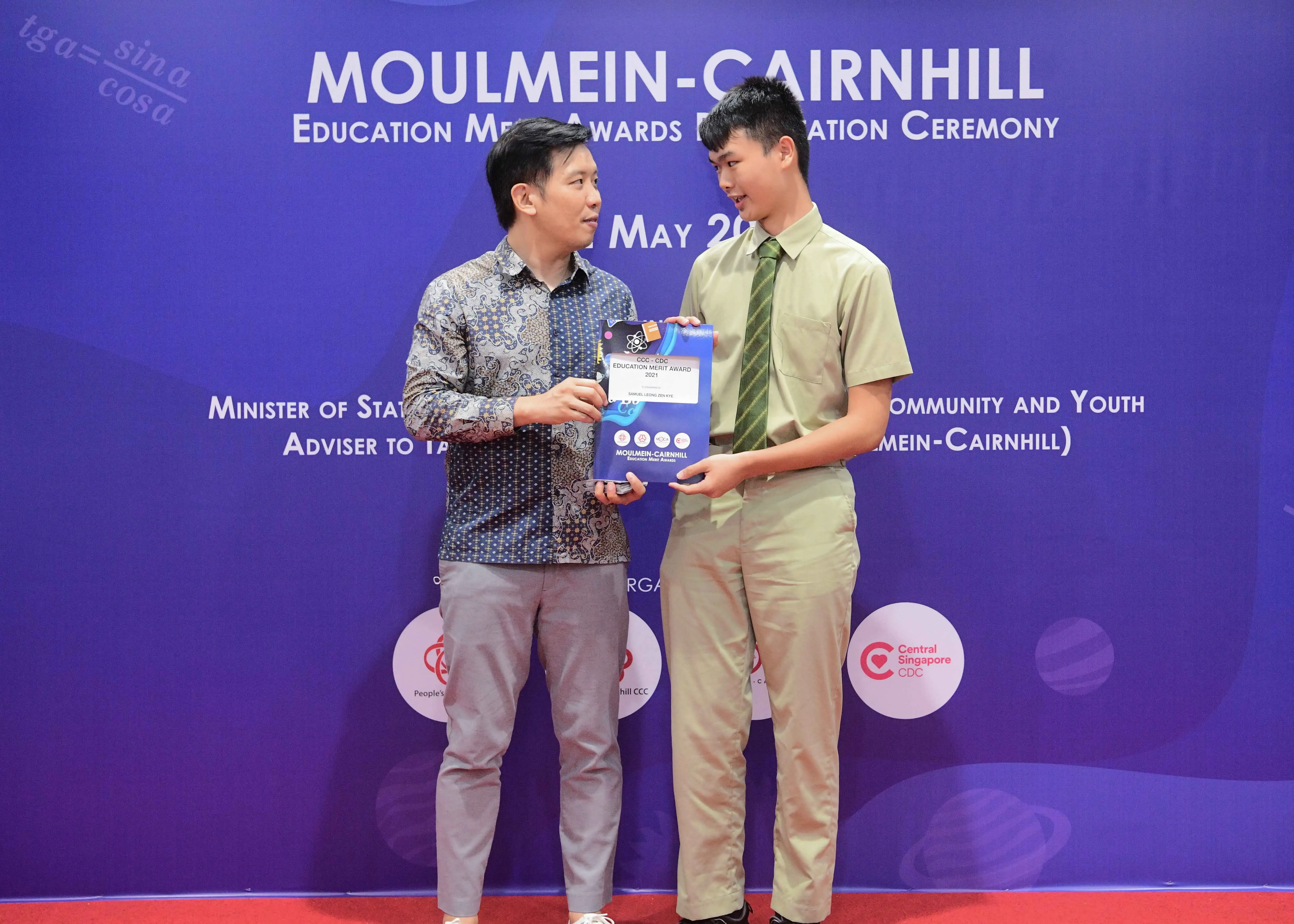 Two men stand before a Moulmein-Cairnhill Education Merit Awards banner, jointly holding an award certificate.
