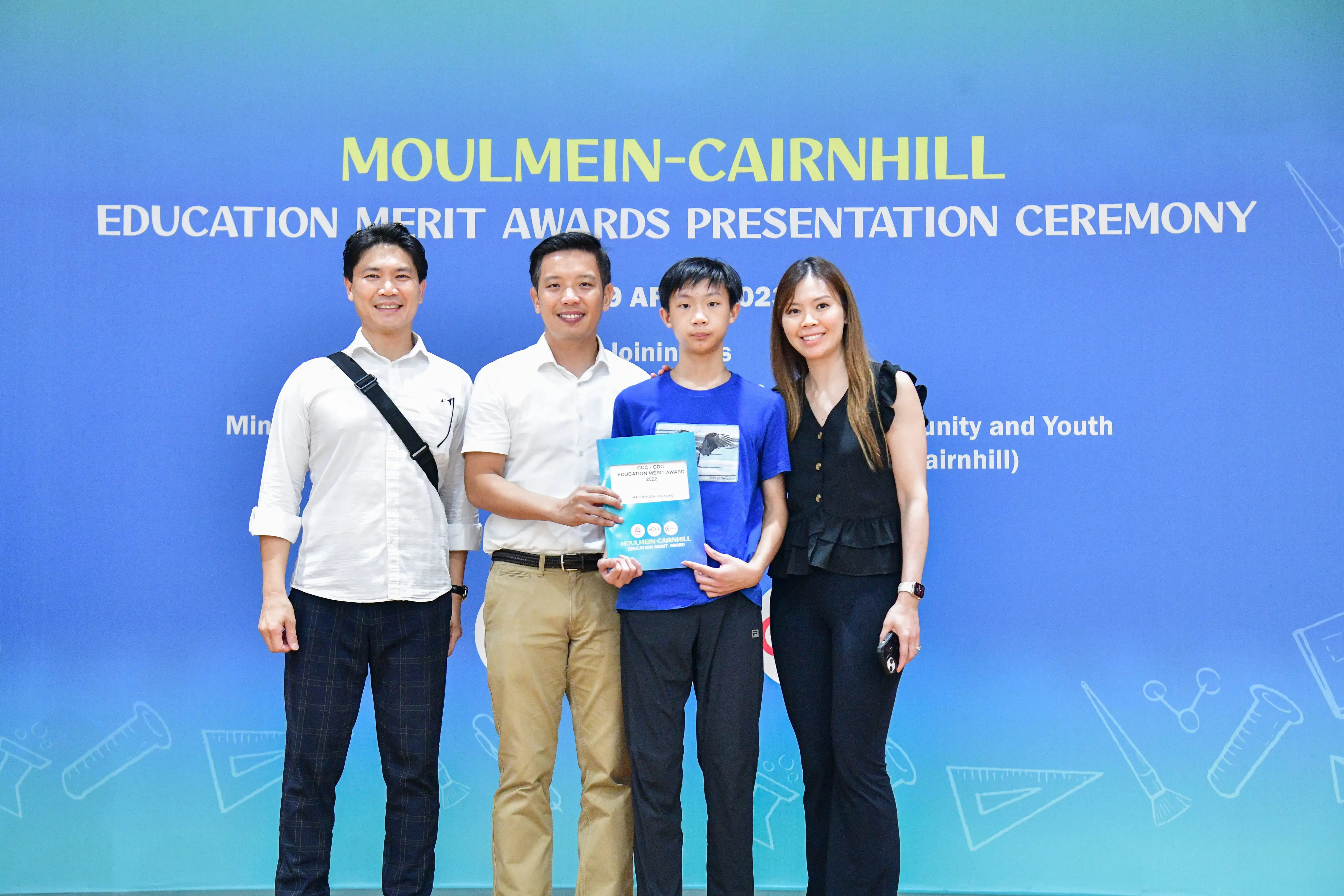 Four people posing in front of a Moulmein-Cairnhill Education Merit Awards Presentation Ceremony banner.