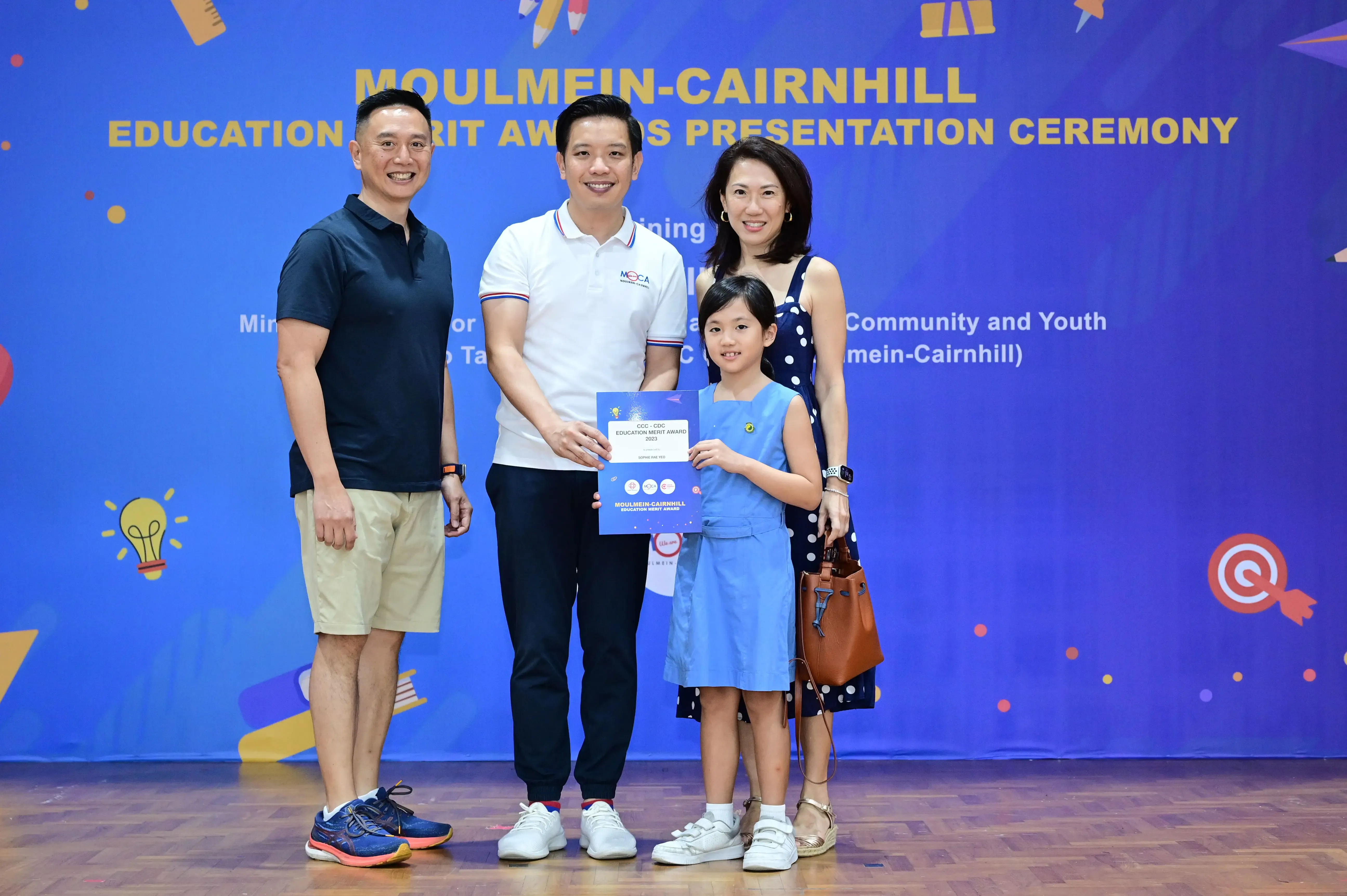 Four people pose at the Moulmein-Cairnhill Education Merit Awards Presentation Ceremony, with a girl holding a certificate.