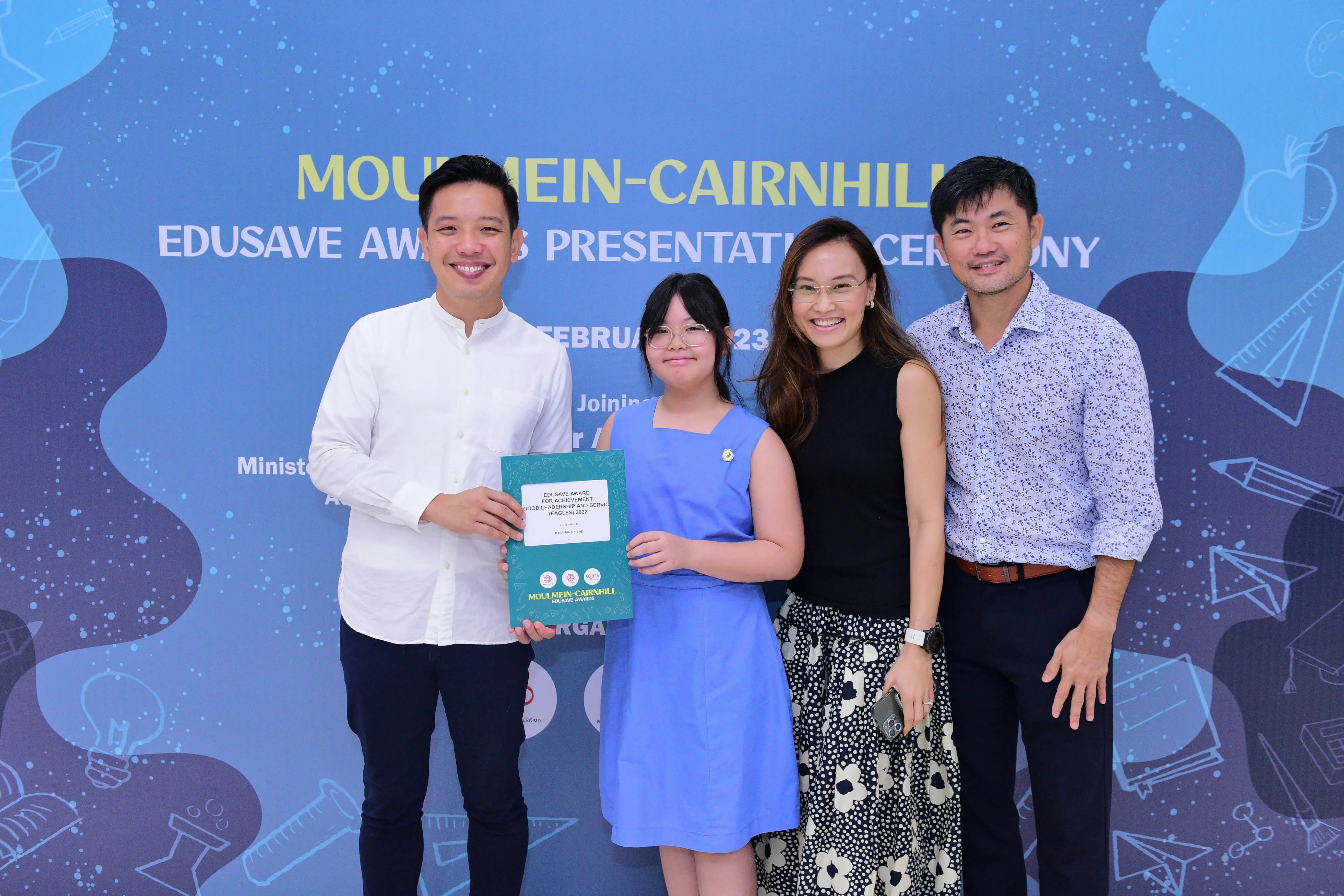 Four people smile and pose at Moulmein-Cairnhill Edusave Awards Presentation Ceremony, girl holds a teal certificate.