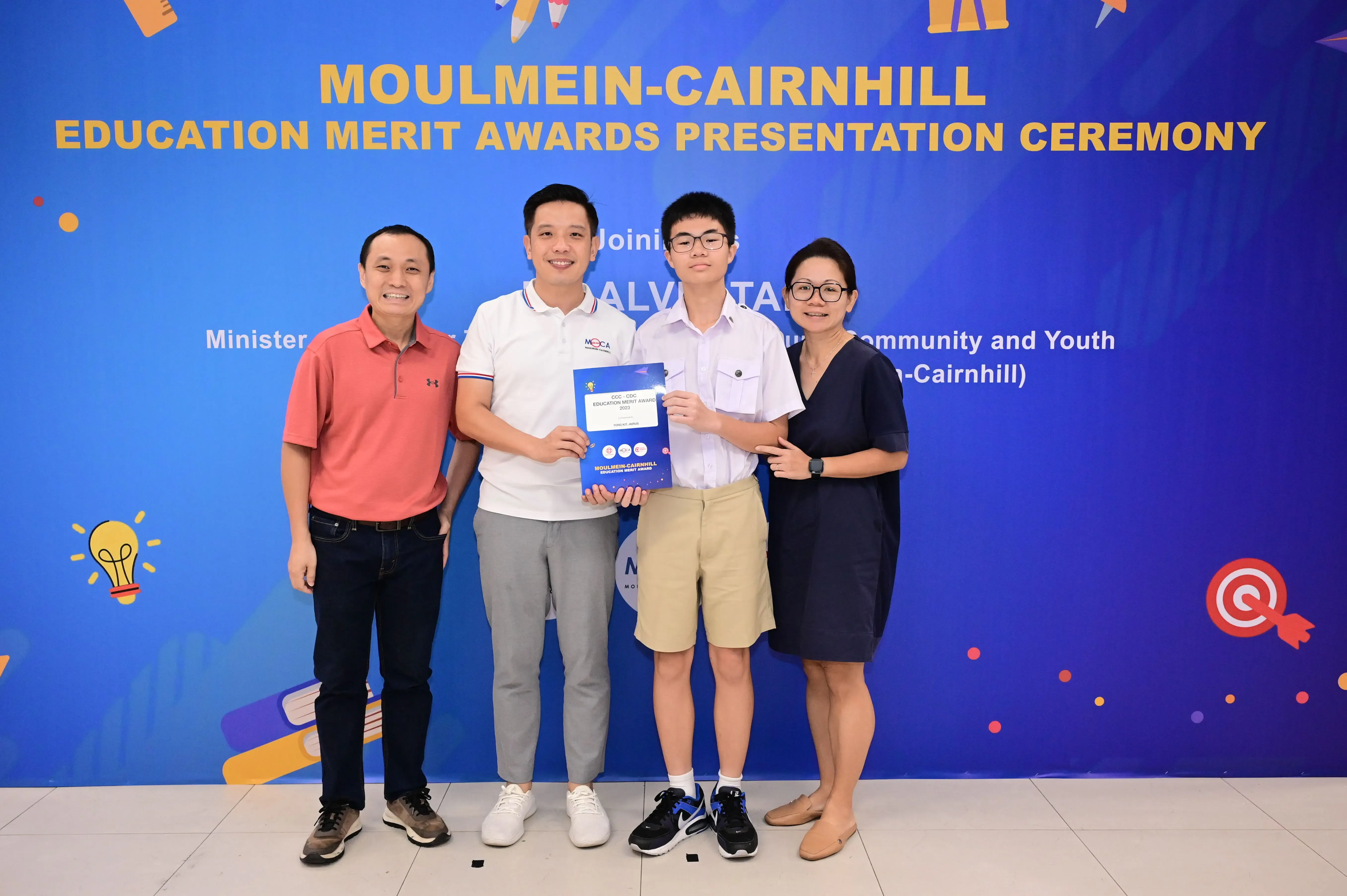 Four people pose at the Moulmein-Cairnhill Education Merit Awards ceremony, with a teen holding a blue certificate.