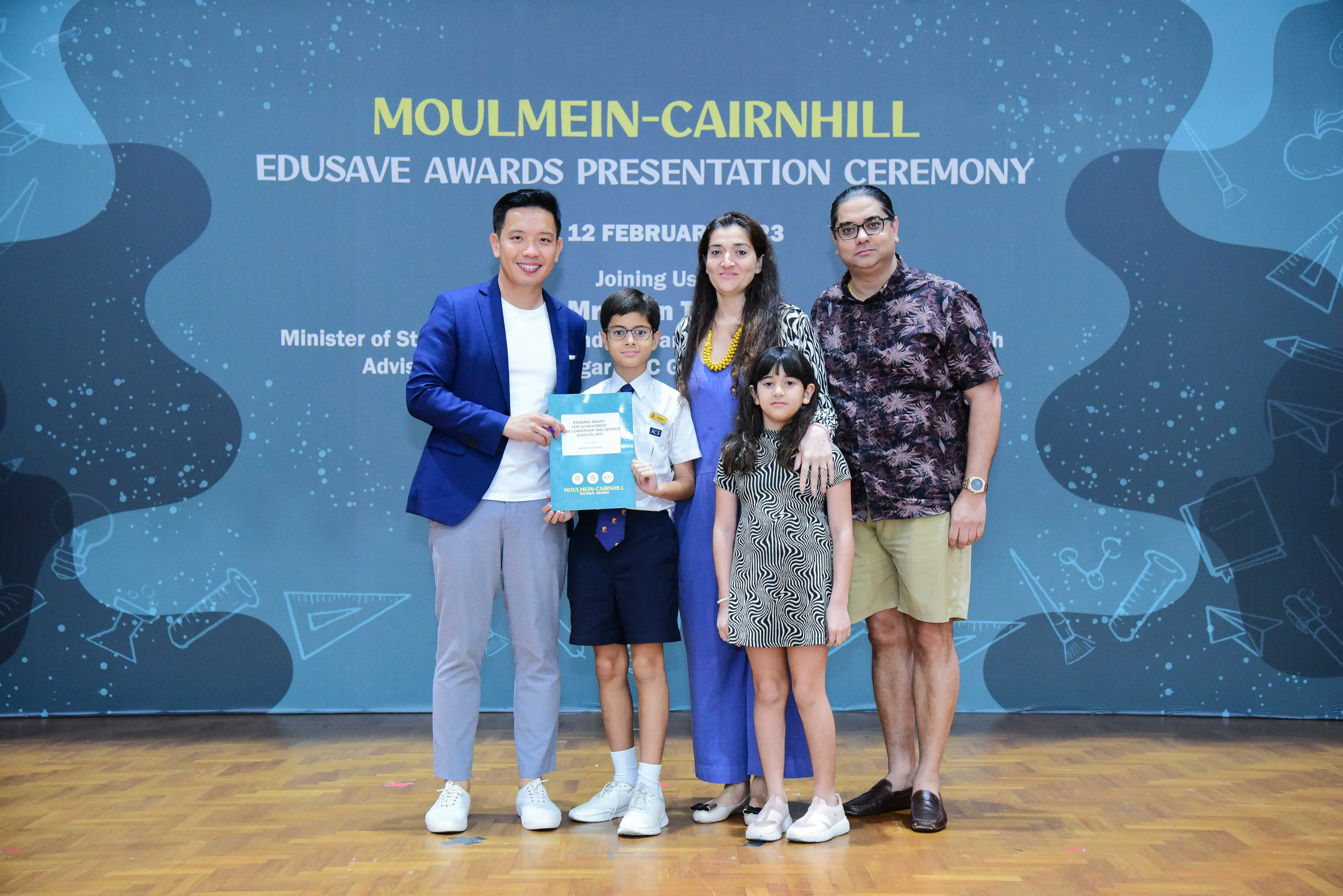 Five people pose at the Moulmein-Cairnhill Edusave Awards Presentation Ceremony, including a boy holding an award certificate.