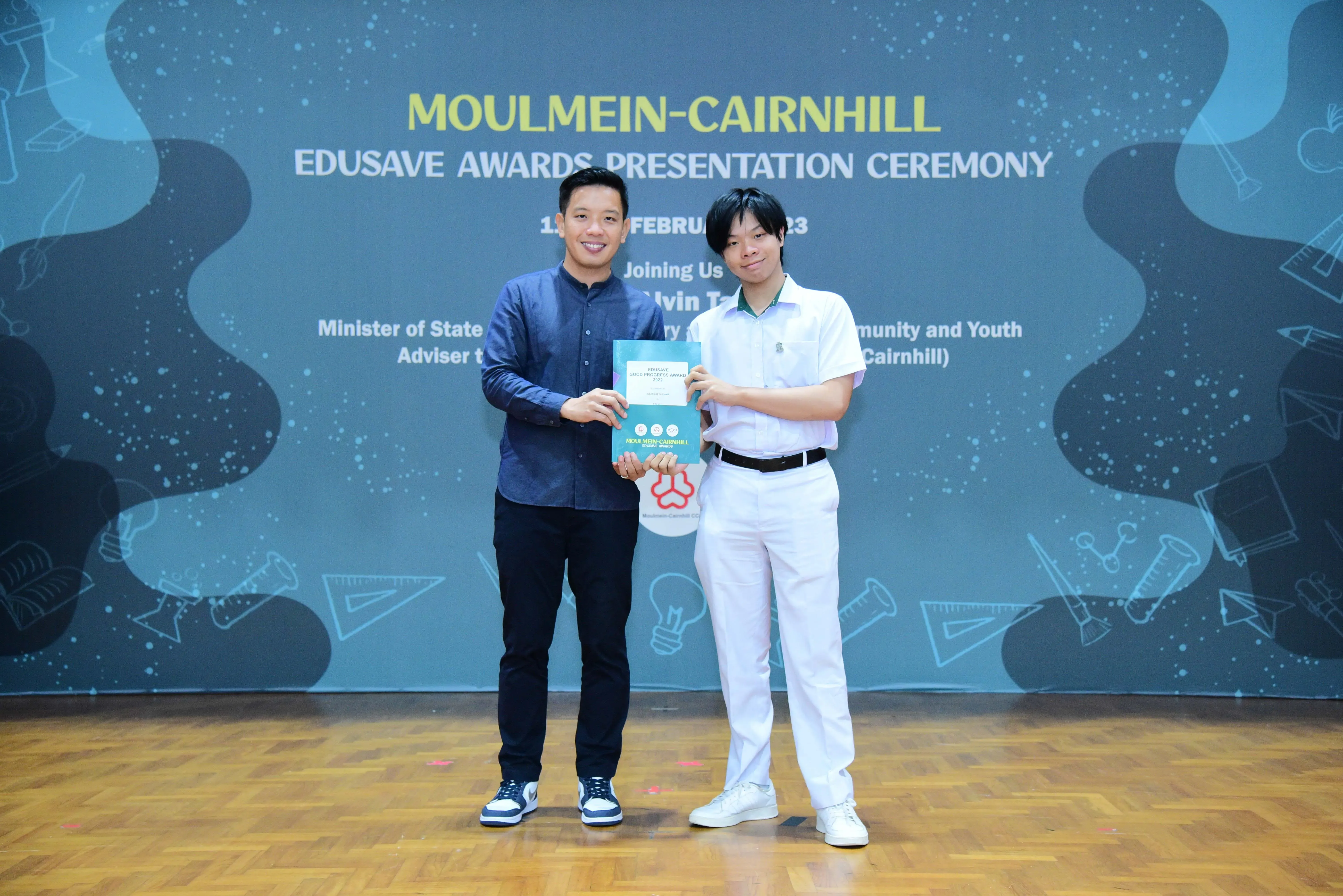 Two men hold a certificate together at the Moulmein-Cairnhill Edusave Awards Presentation Ceremony backdrop.
