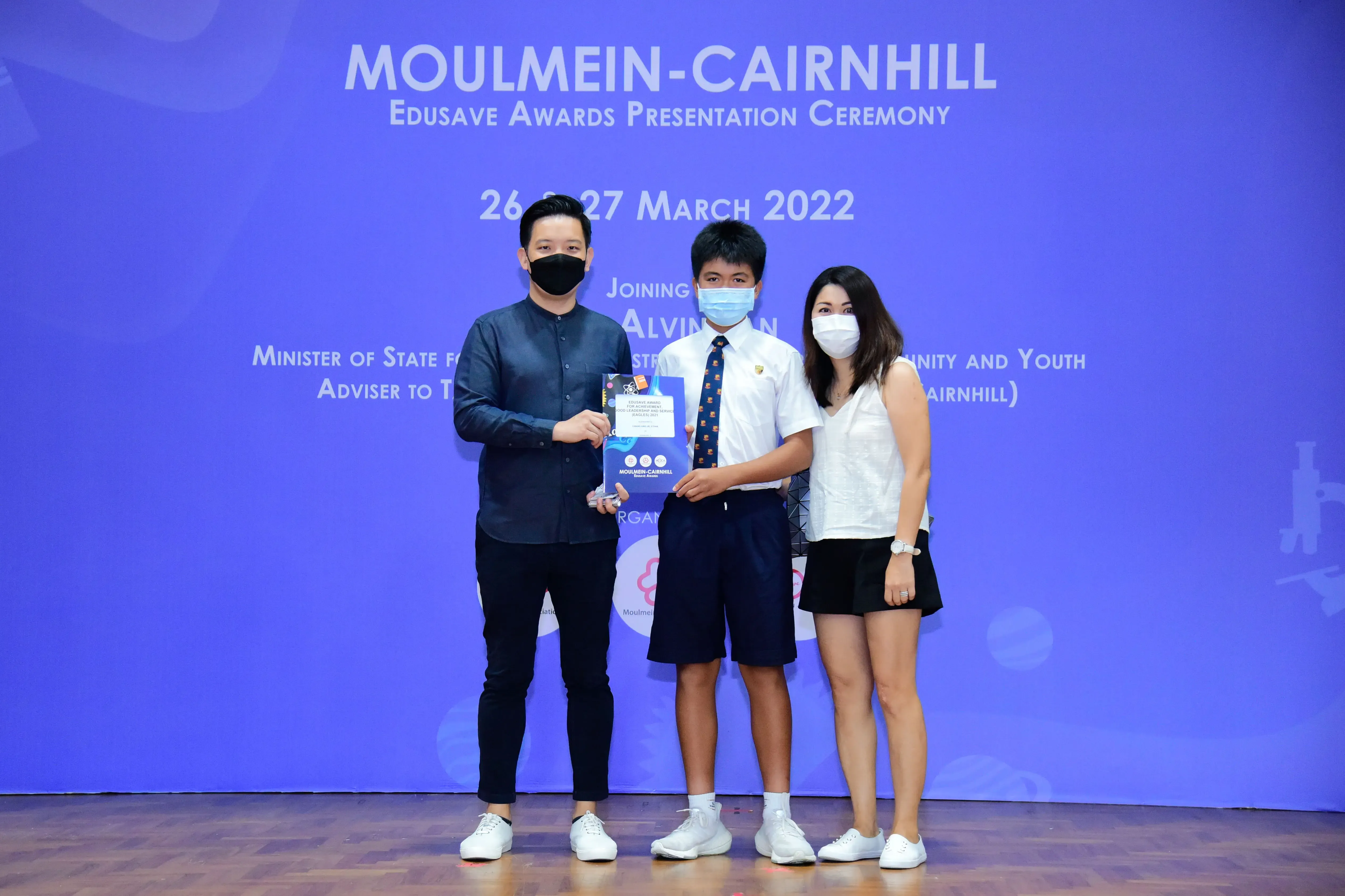 Three masked people pose at the Moulmein-Cairnhill Edusave Awards Ceremony on 26-27 March 2022, holding a trophy.