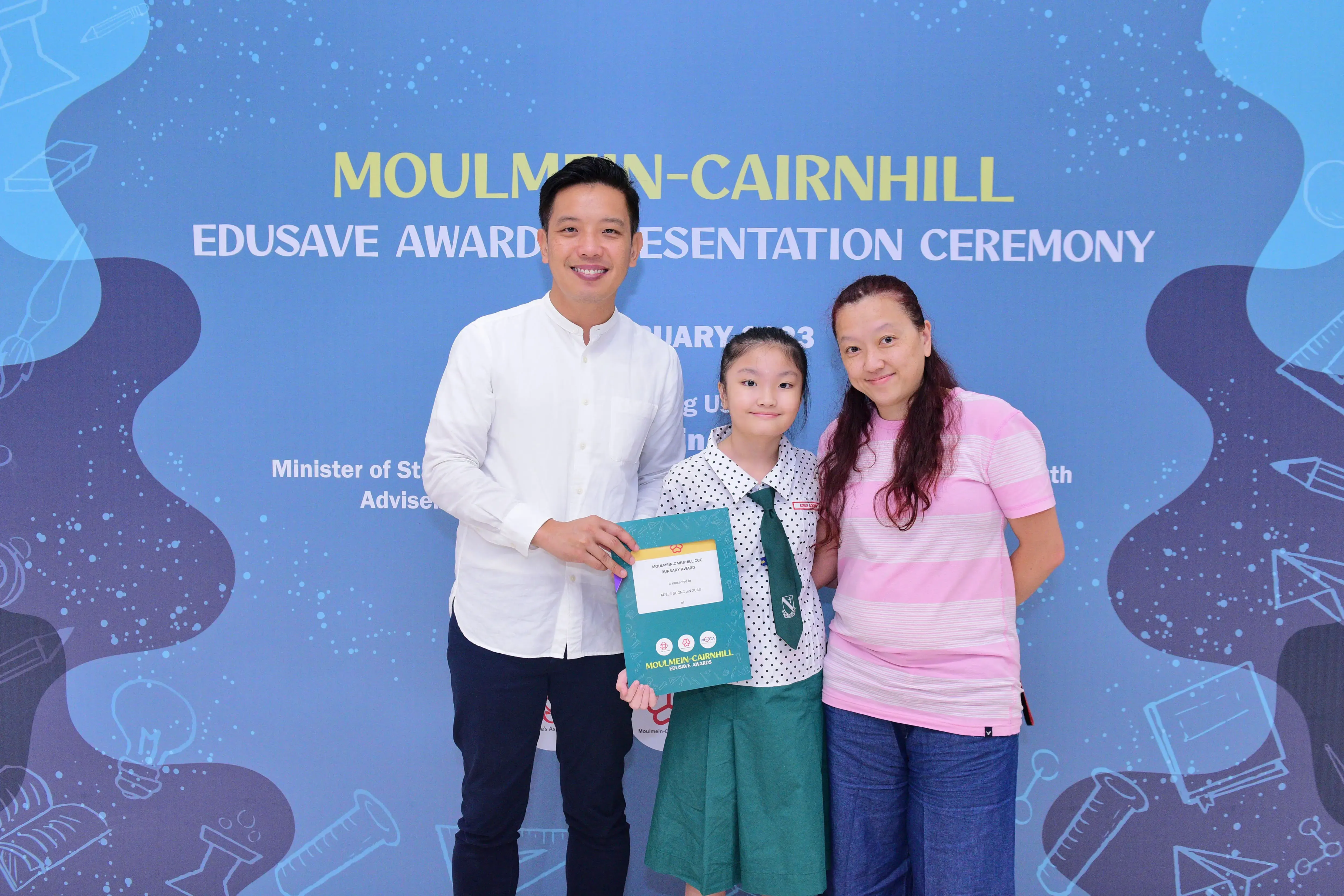 A man, a young girl holding a certificate, and a woman pose at the Moulmein-Cairnhill Edusave Awards Ceremony.