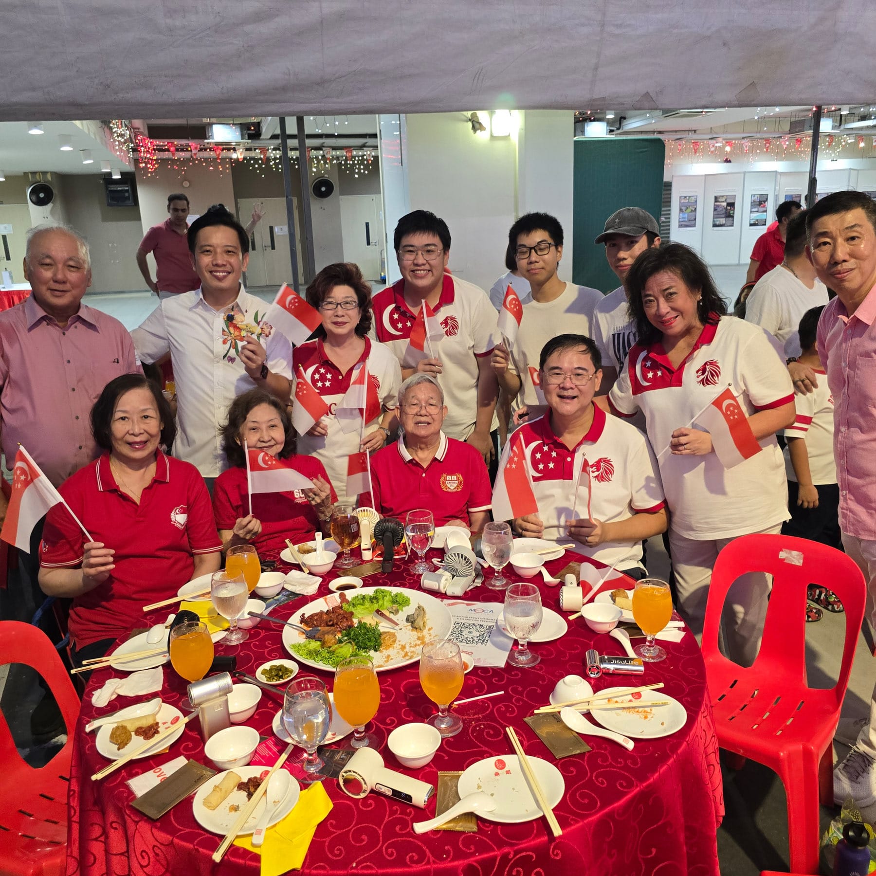 Group of people in red and white outfits holding Singapore flags, seated and standing around a round dining table with food.