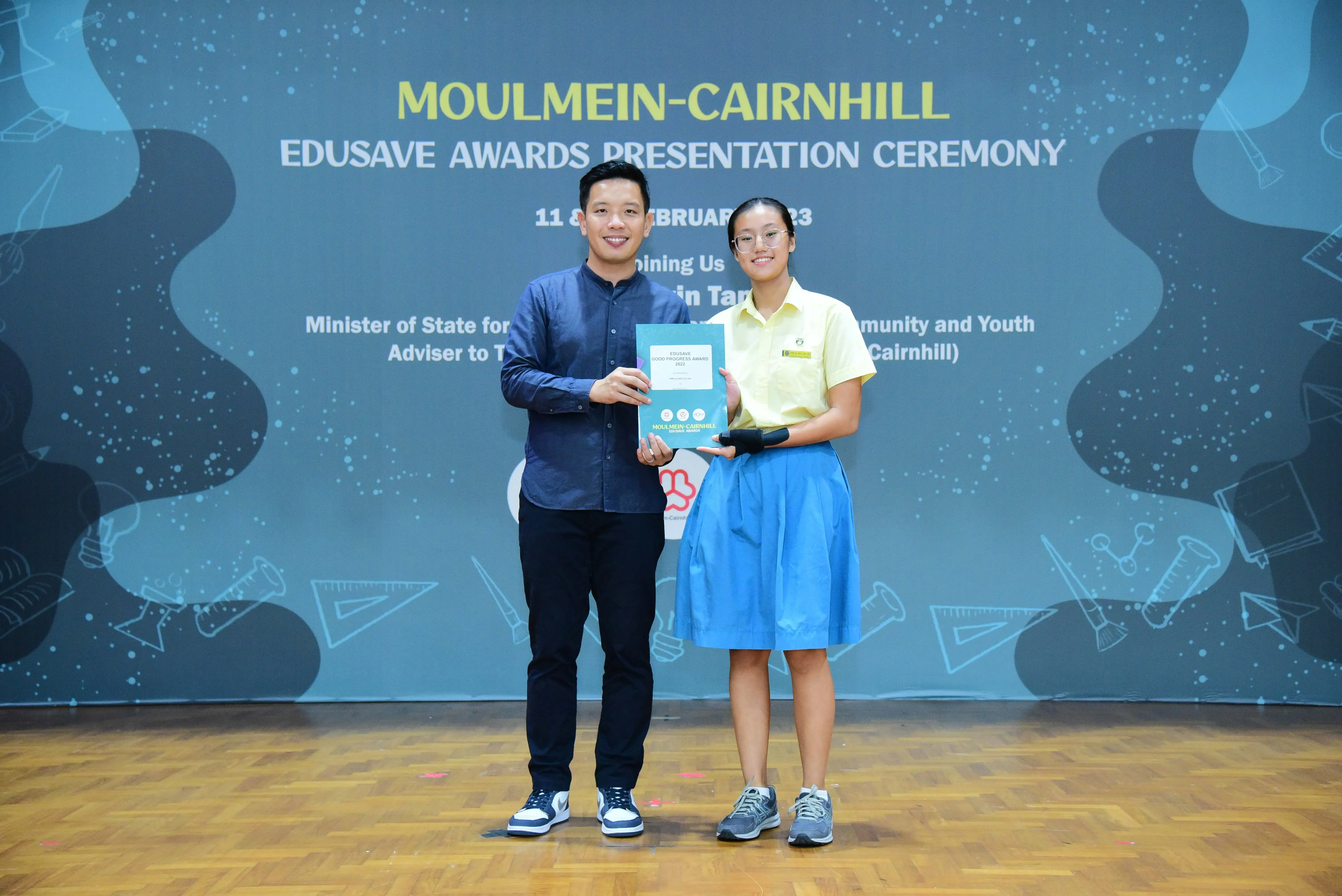 Two people hold a certificate together at the Moulmein-Cairnhill Edusave Awards Presentation Ceremony backdrop.