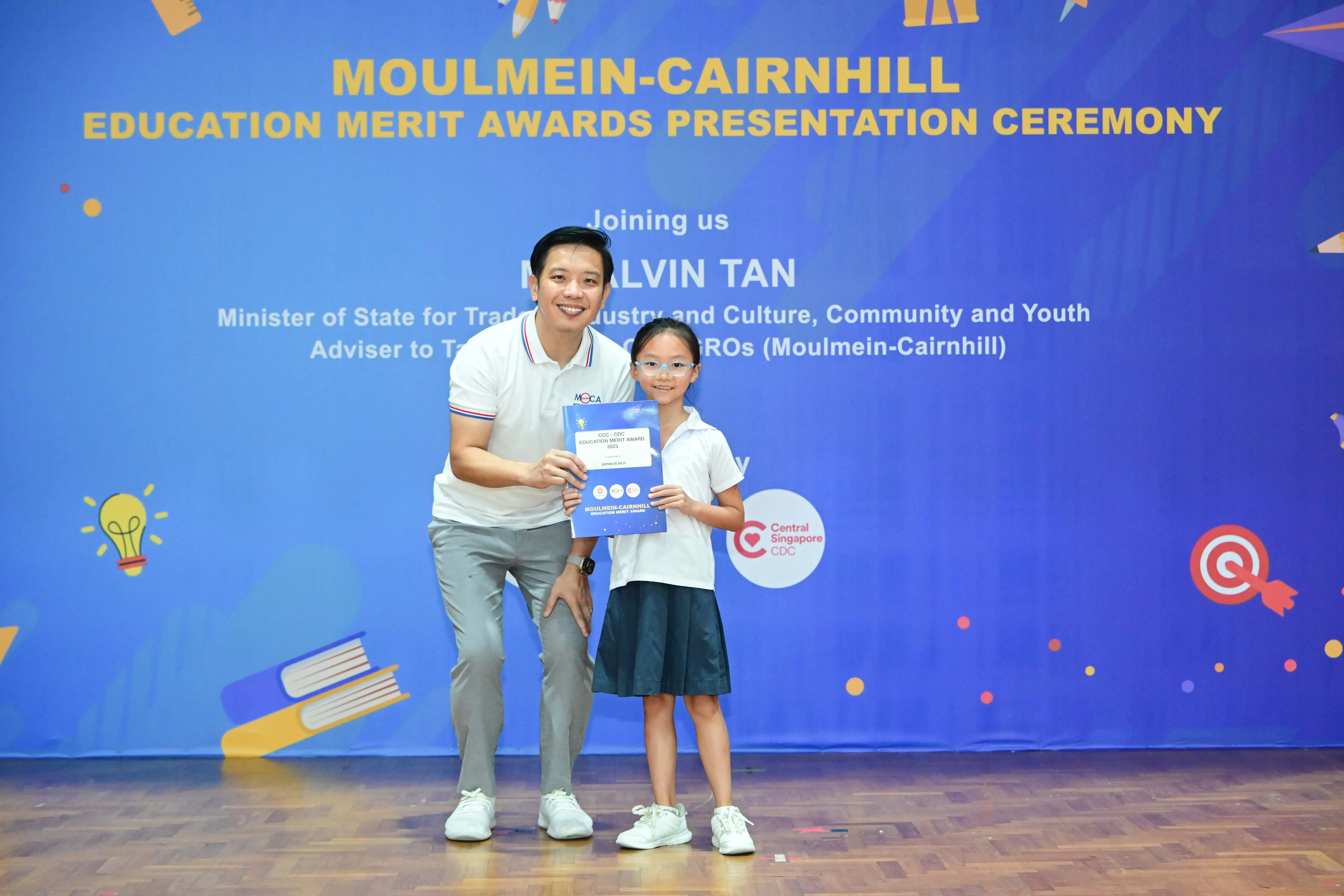 A man and a young girl hold a certificate in front of a Moulmein-Cairnhill Education Merit Awards banner.