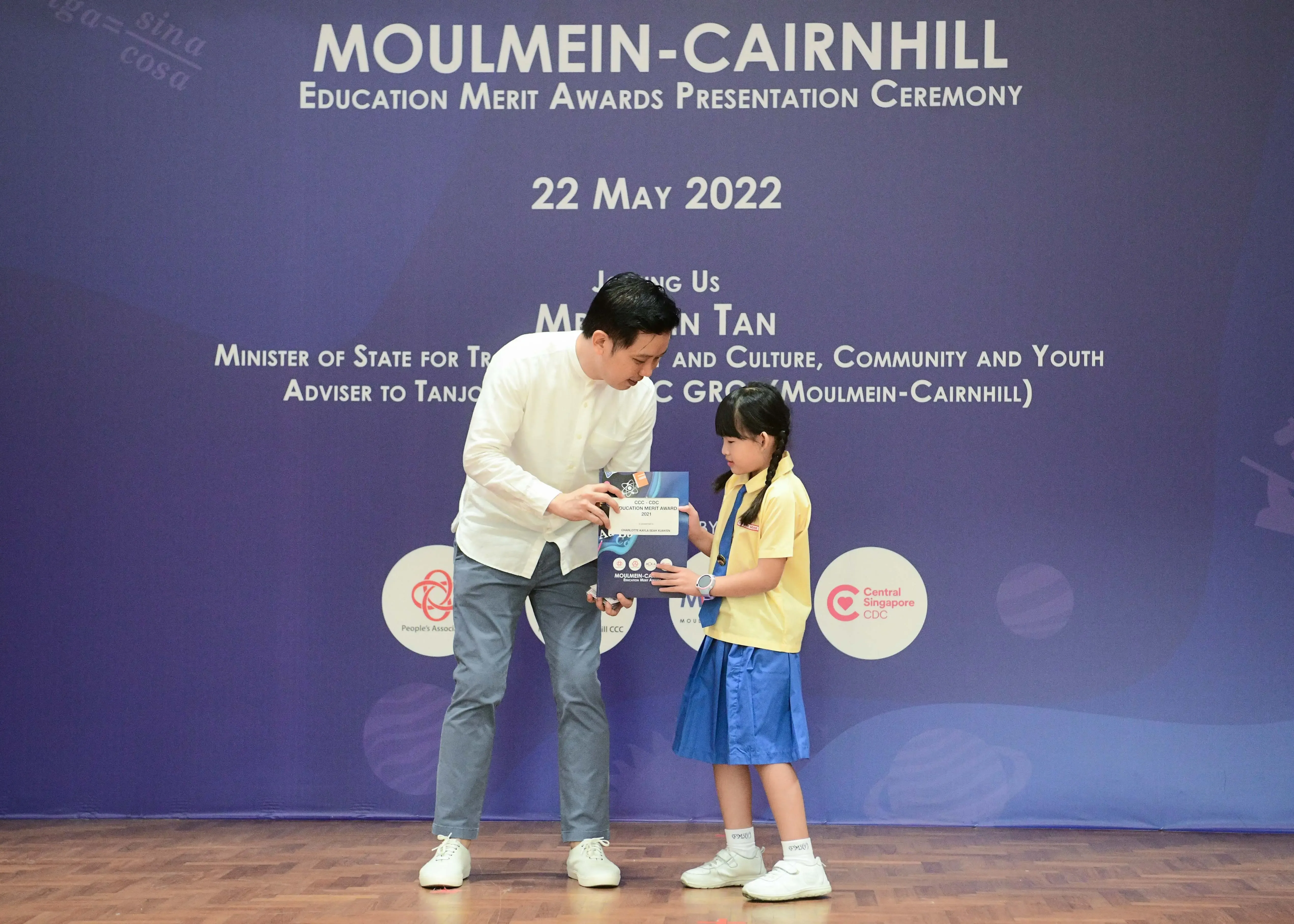 A man in white shirt presents an award to a young girl in yellow uniform at the Moulmein-Cairnhill Education Merit Awards 2022.