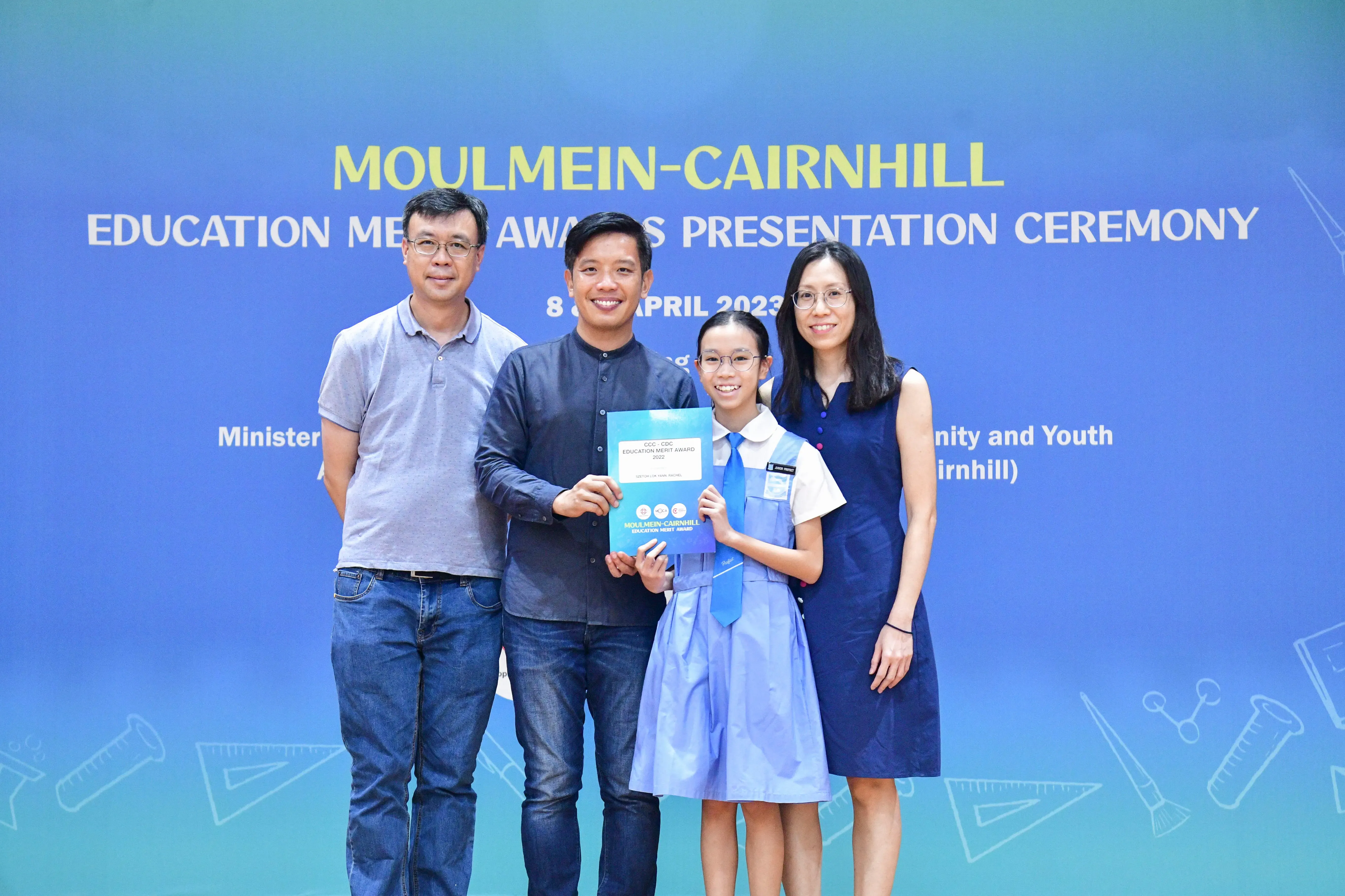 Four people pose before a Moulmein-Cairnhill Education Merit Awards Presentation Ceremony banner, with a girl holding an award.