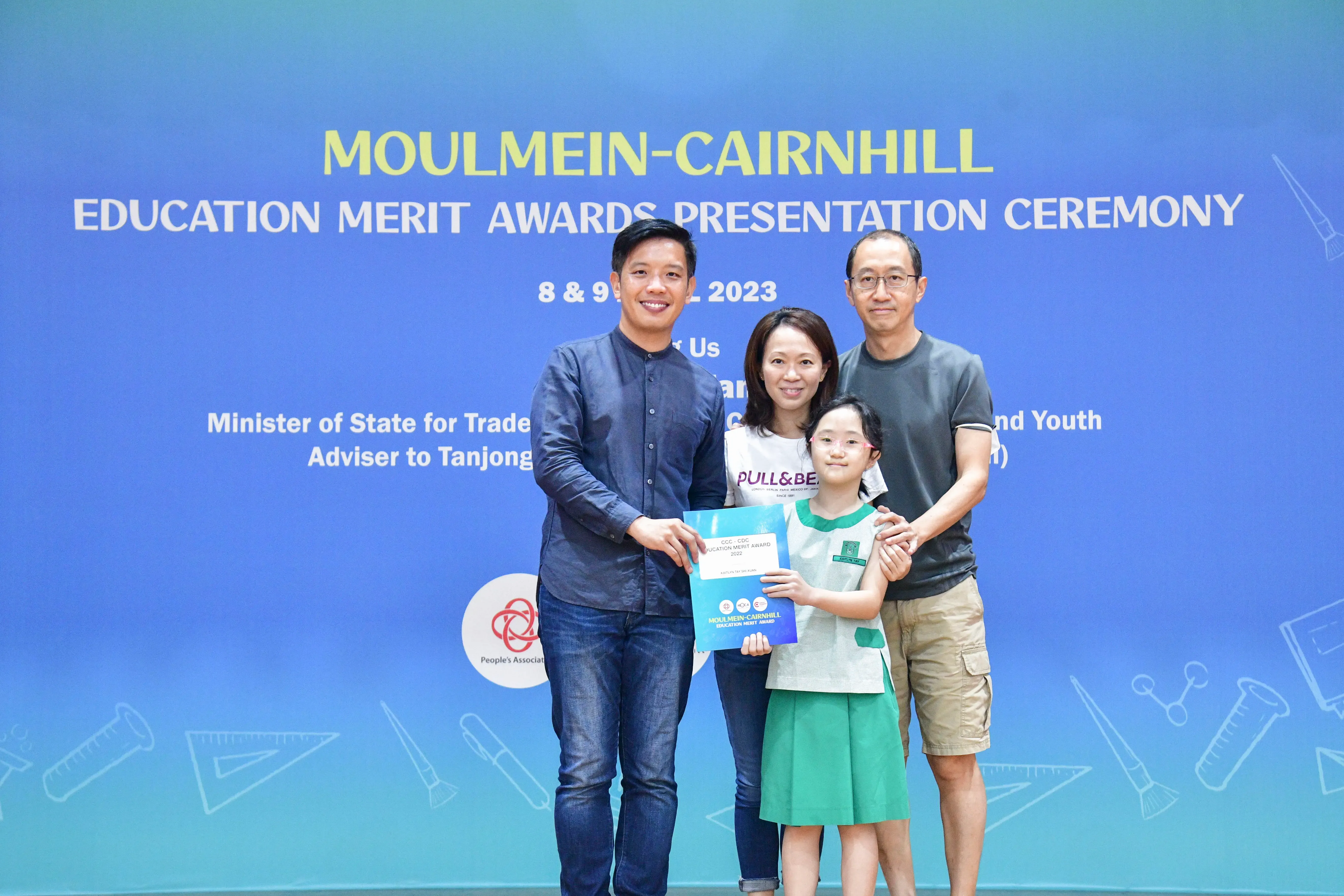 Three adults and a girl holding award certificates in front of a Moulmein-Cairnhill Education Merit Awards 2023 banner.