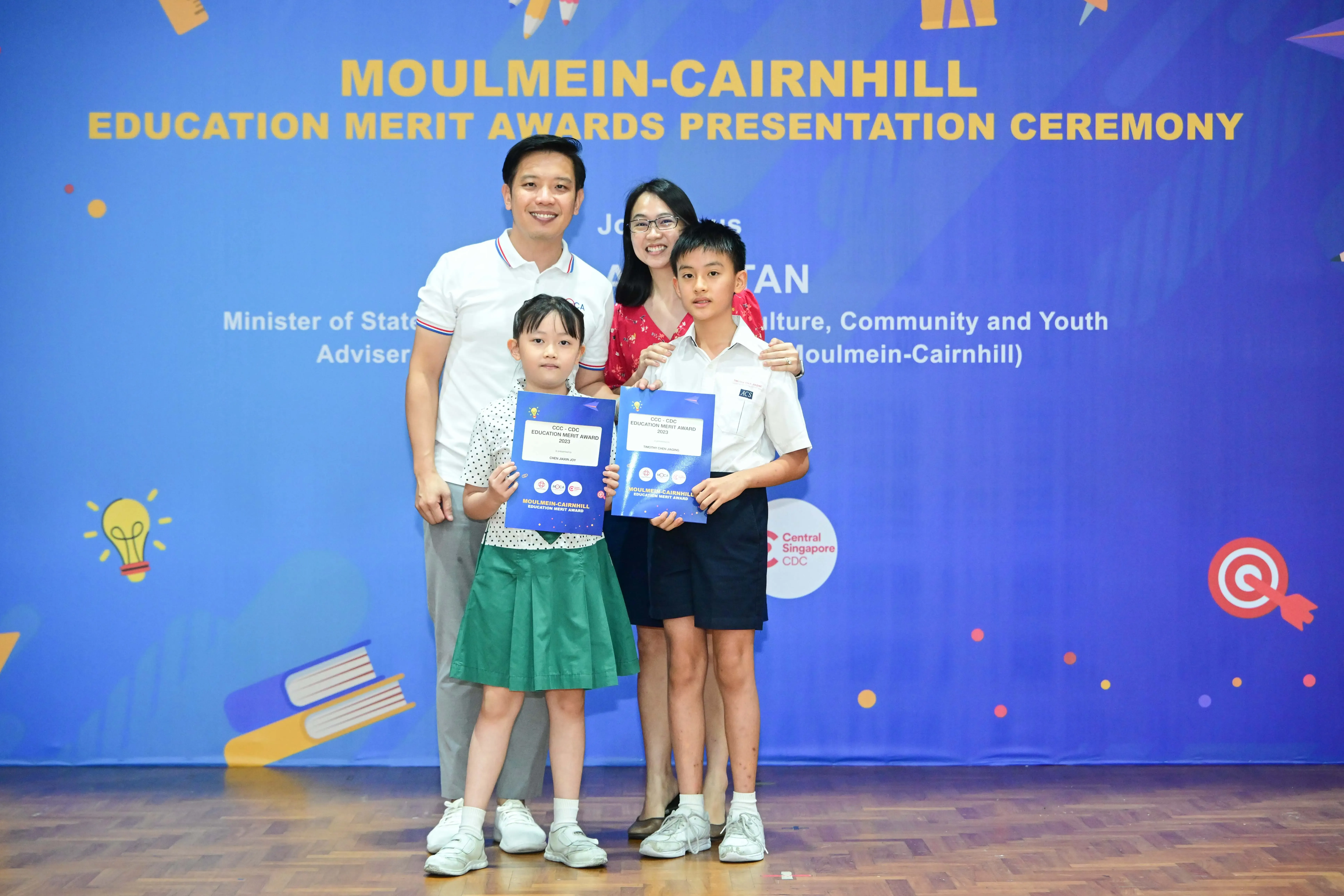 Two adults and two children pose holding award certificates at the Moulmein-Cairnhill Education Merit Awards ceremony.