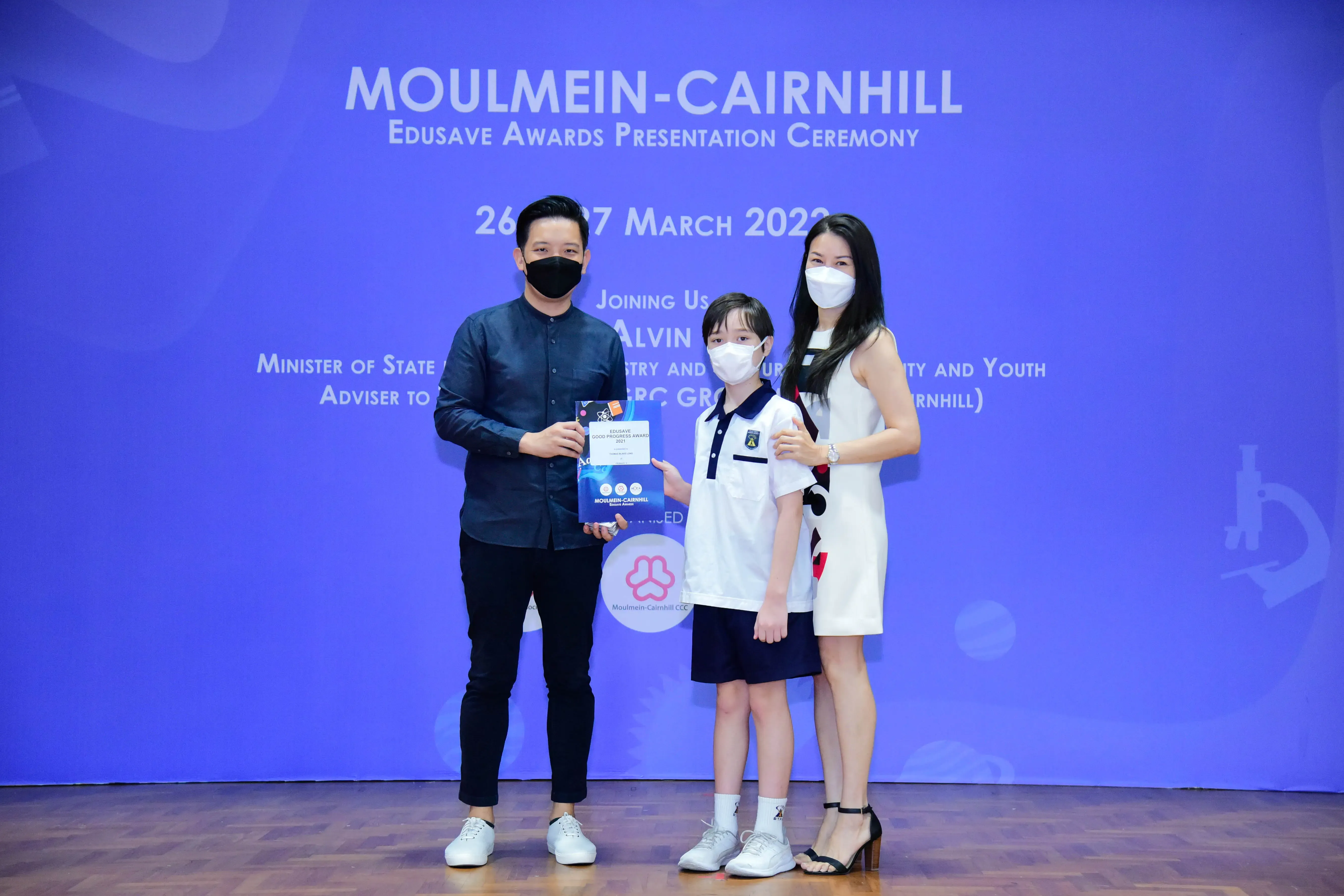 Two masked adults and a masked child in school uniform hold an award at the Moulmein-Cairnhill Edusave ceremony.