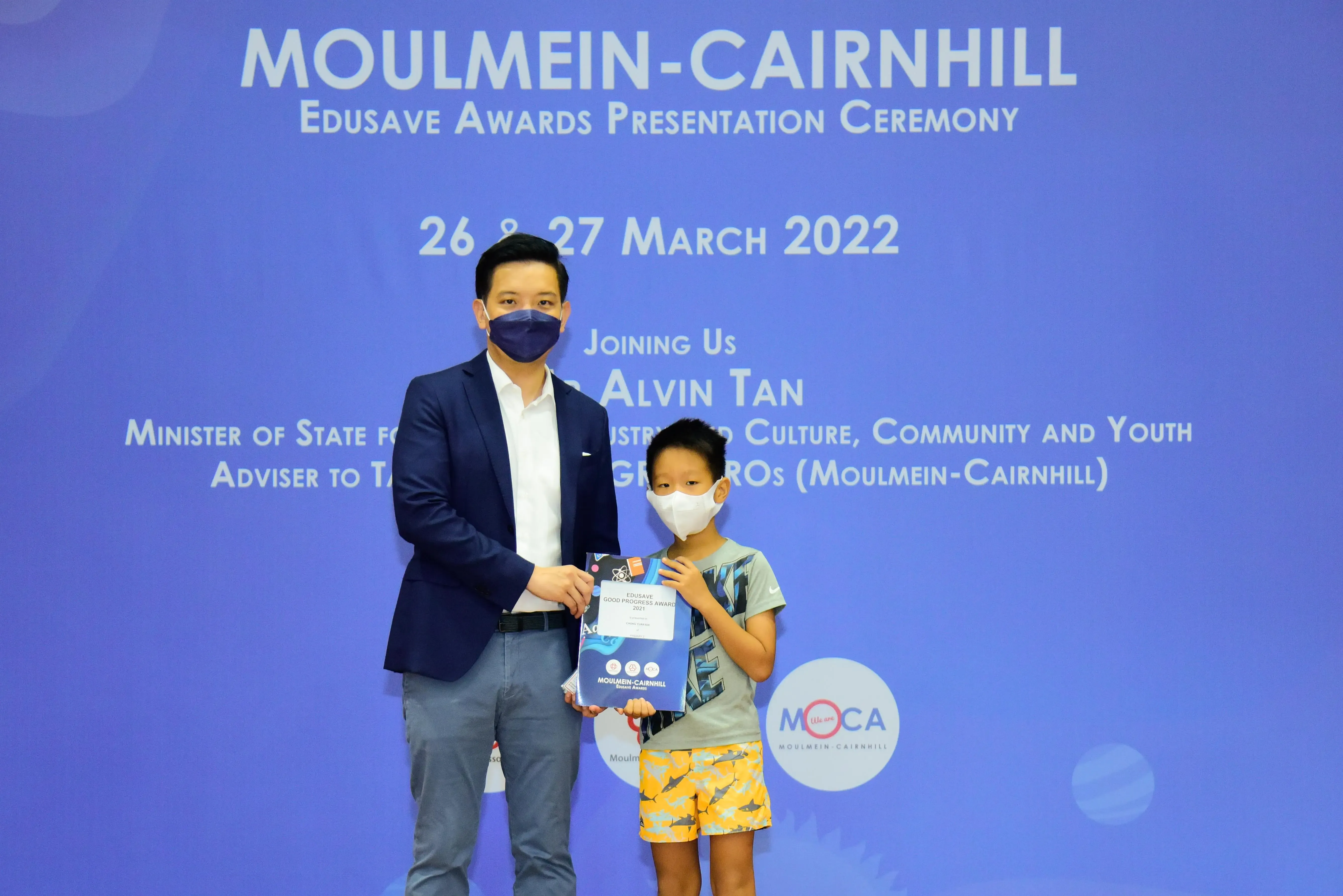 An adult and a young boy wearing masks hold a certificate at the Moulmein-Cairnhill Edusave Awards Ceremony 2022.