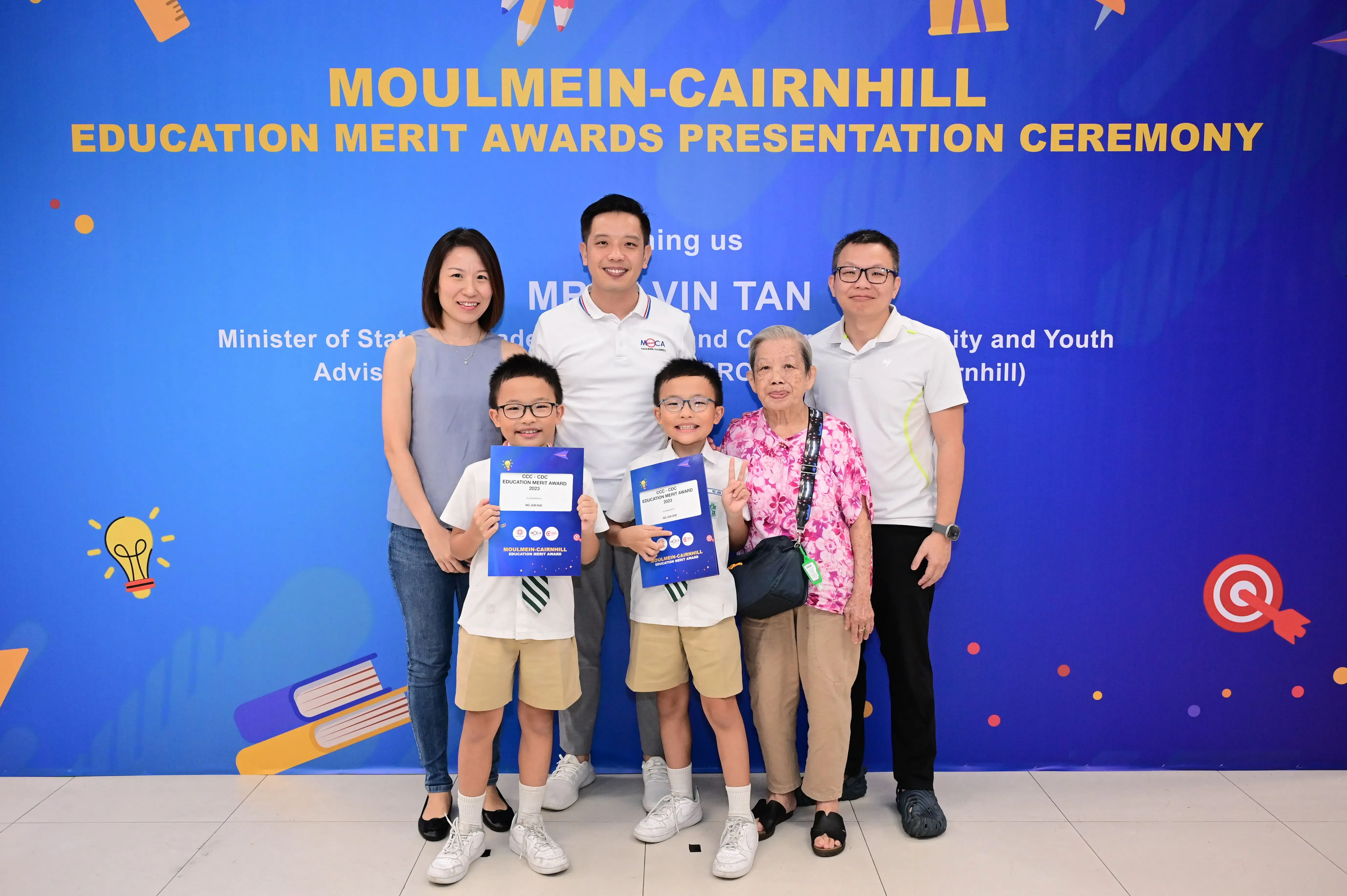 Six people pose before a Moulmein-Cairnhill Education Merit Awards banner; two boys hold blue award certificates.