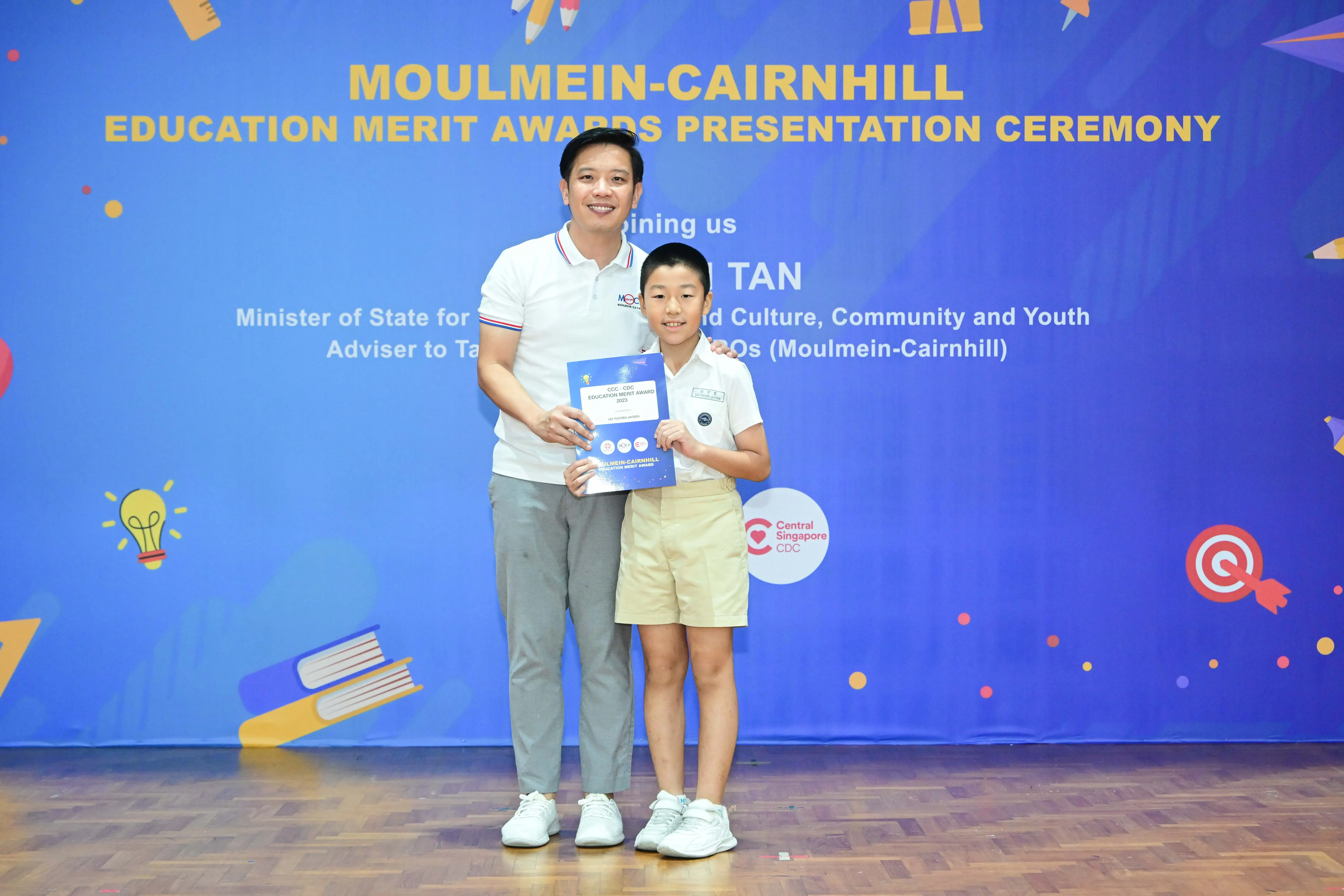 A man and a young boy hold a certificate in front of the Moulmein-Cairnhill Education Merit Awards banner.
