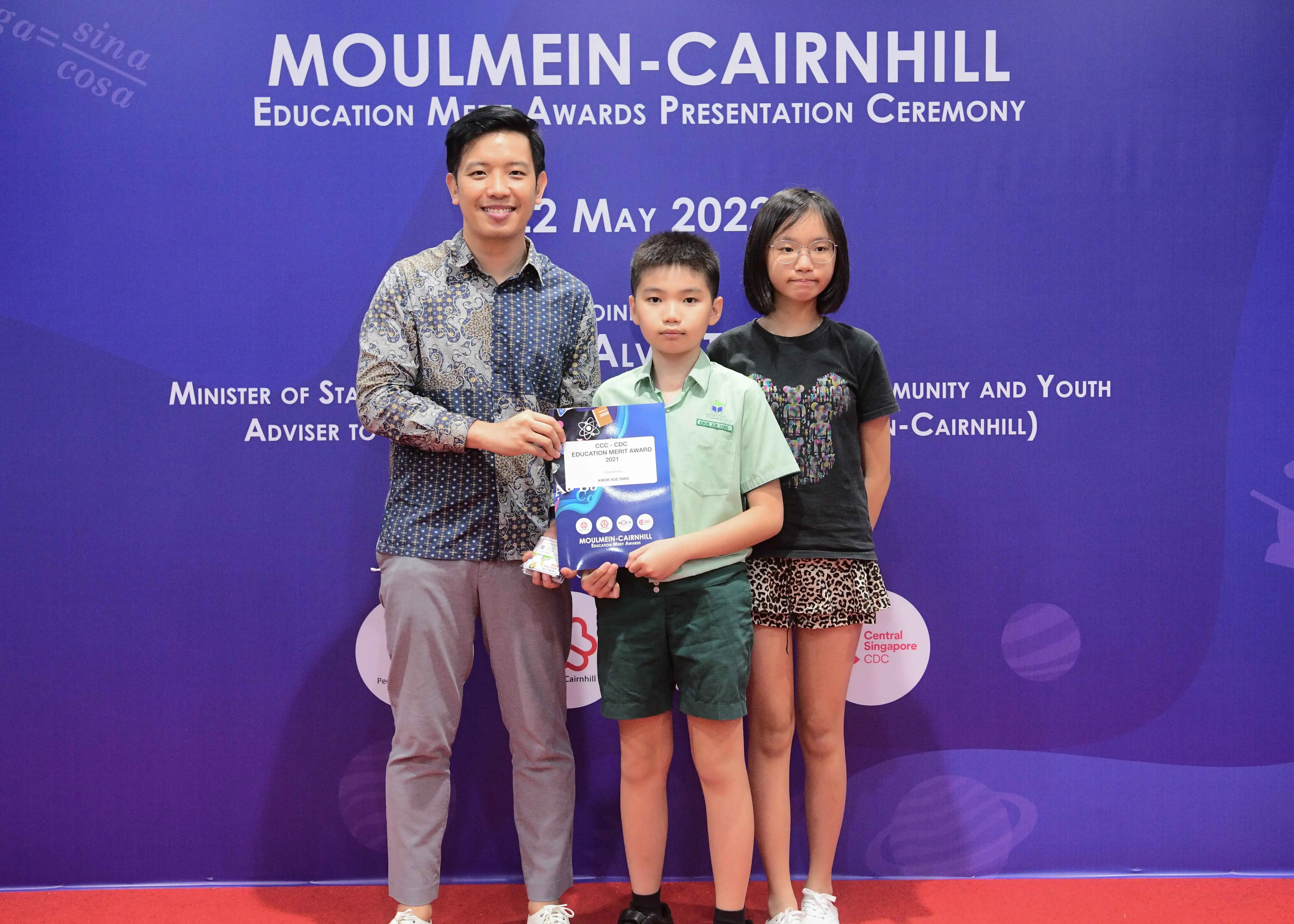 Three people pose at the Moulmein-Cairnhill Education Merit Awards Presentation Ceremony, with a boy holding an award folder.