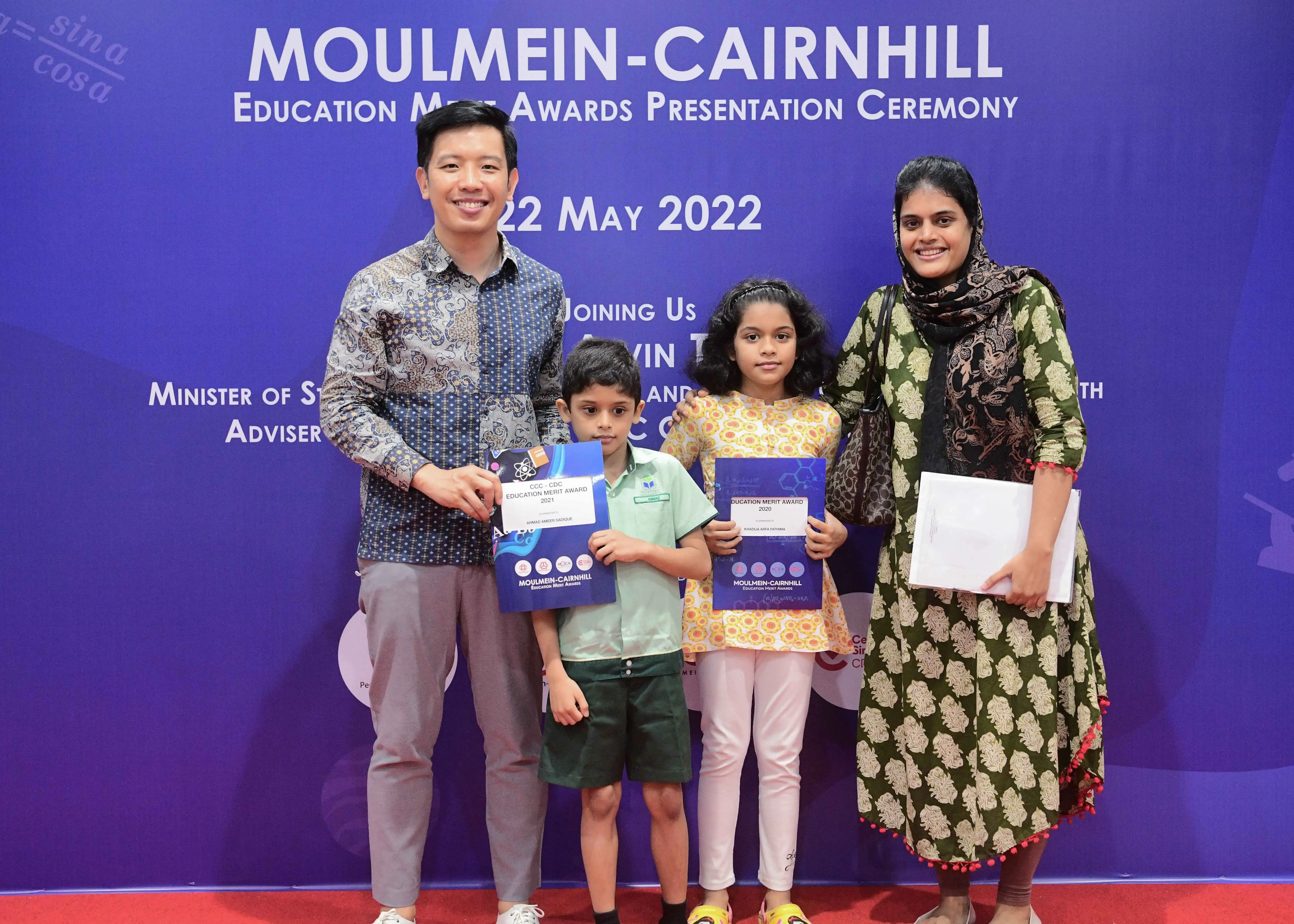 Two adults and two children pose holding merit award certificates at the Moulmein-Cairnhill Education Awards Ceremony, May 2022.