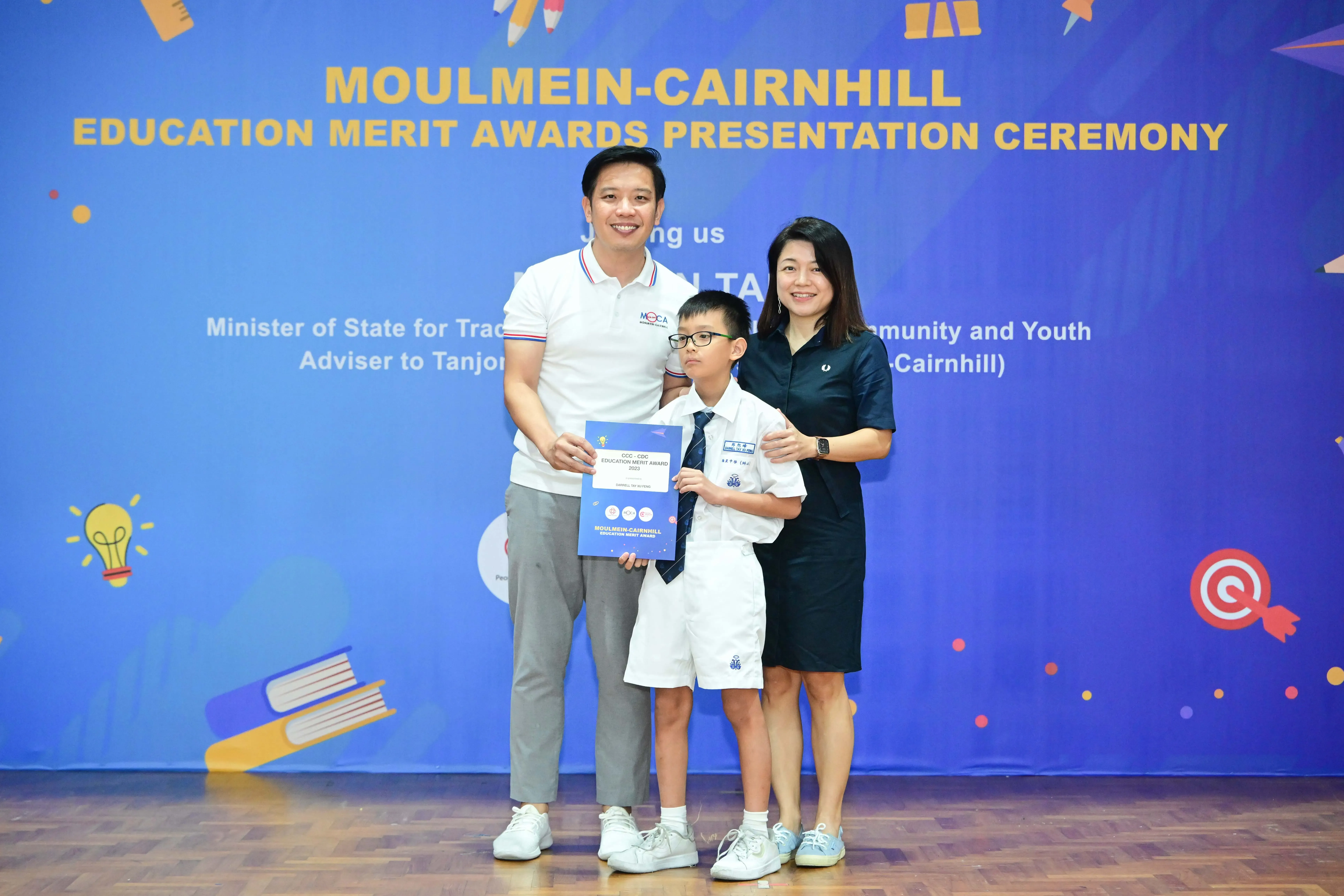 Two adults and a boy in school uniform holding a certificate at Moulmein-Cairnhill Education Merit Awards ceremony.