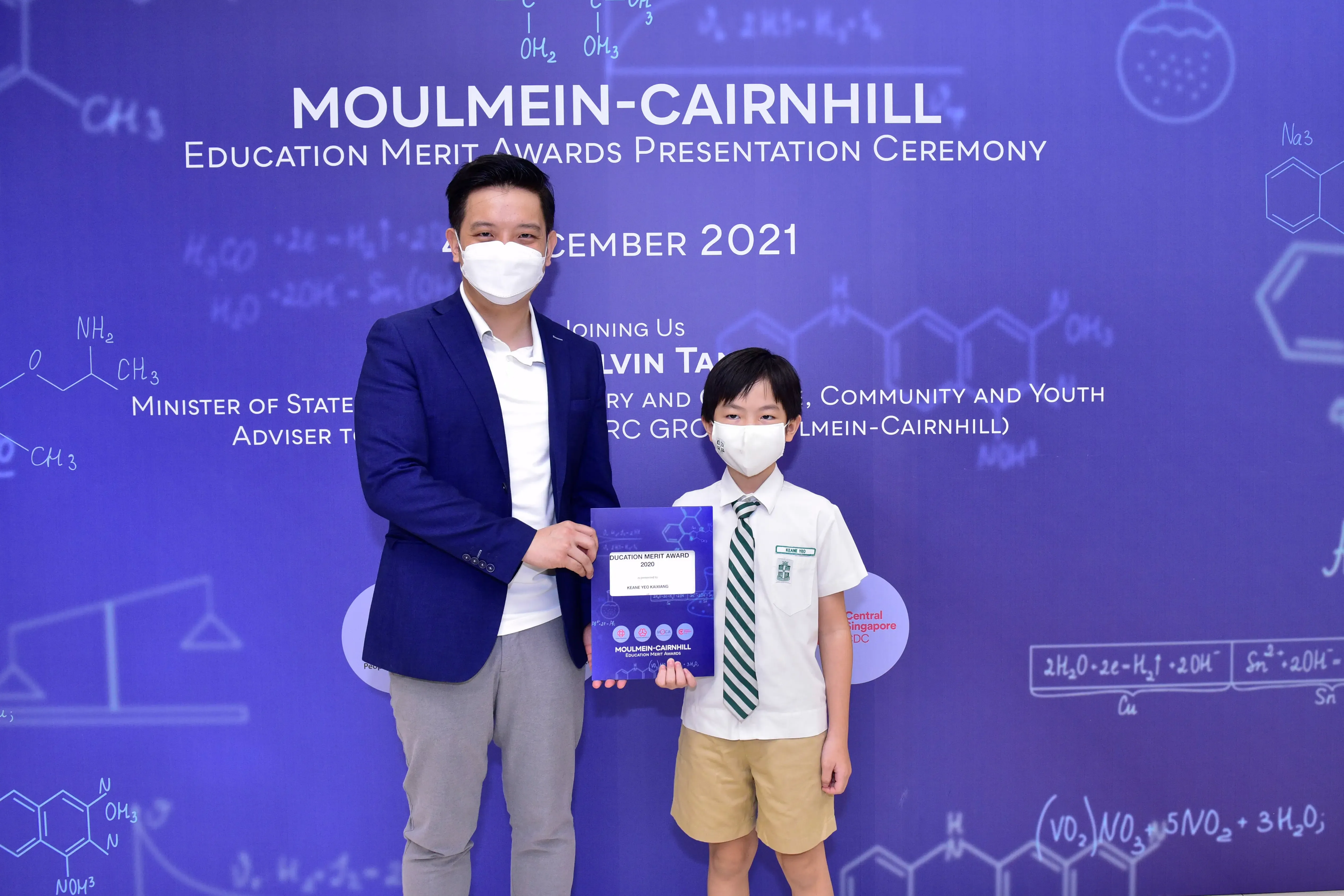 A masked adult and a young boy in school uniform hold an award certificate at the Moulmein-Cairnhill Education Merit Awards 2021.