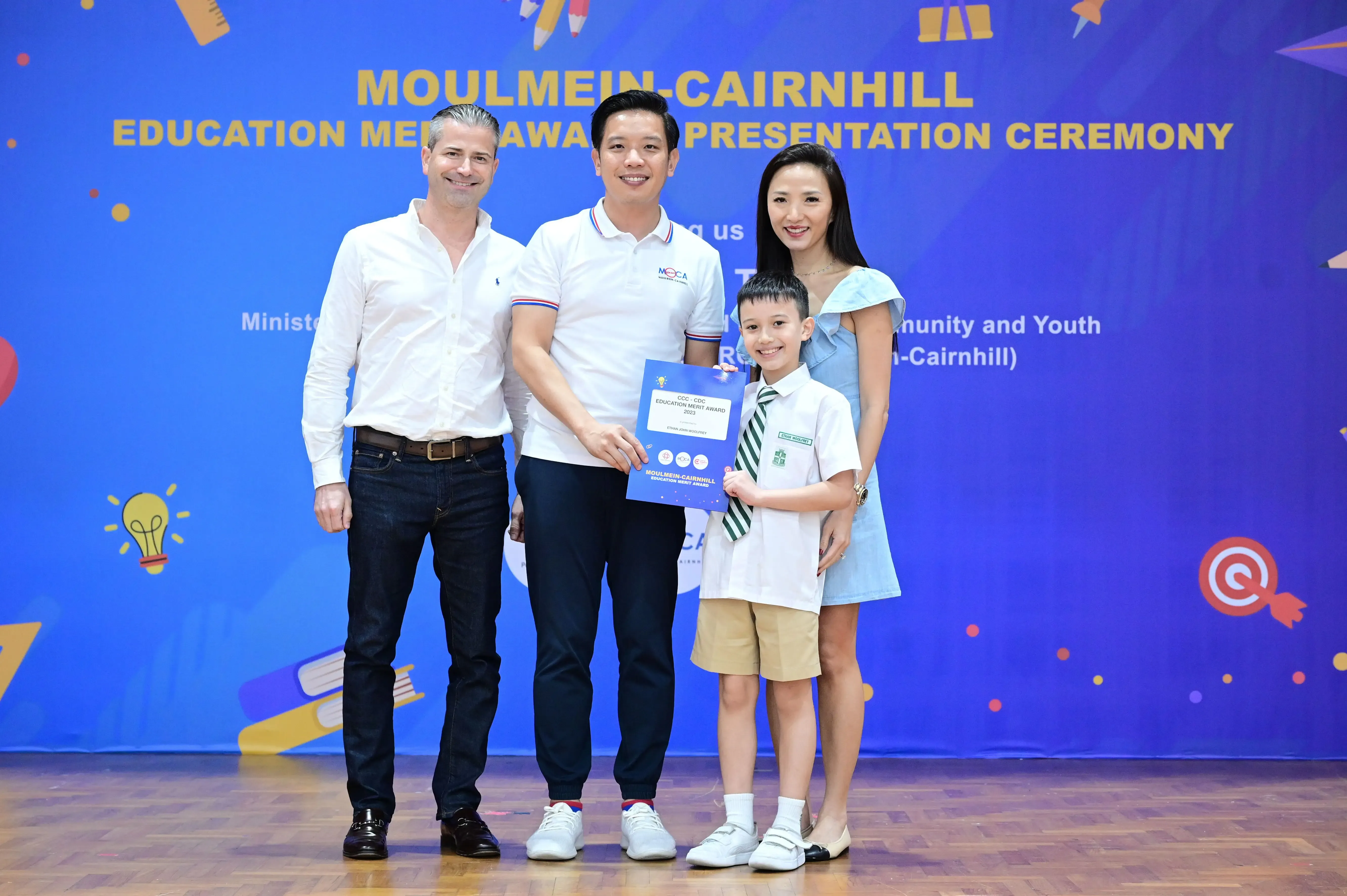Three adults and a boy in school uniform pose with a certificate at the Moulmein-Cairnhill Education Merit Award ceremony.