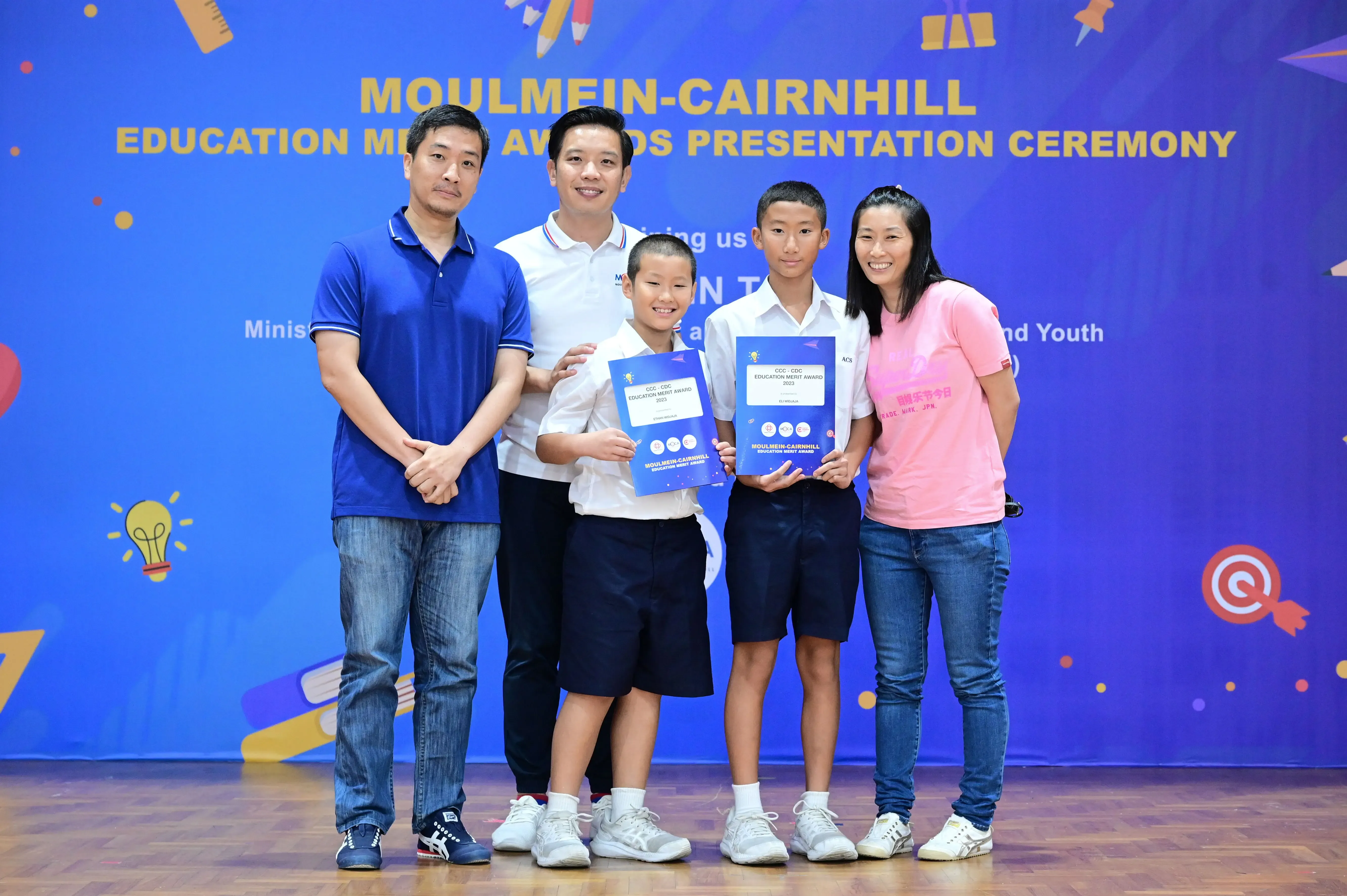 Five people pose at the Moulmein-Cairnhill Education Merit Awards ceremony, two boys holding blue award certificates.
