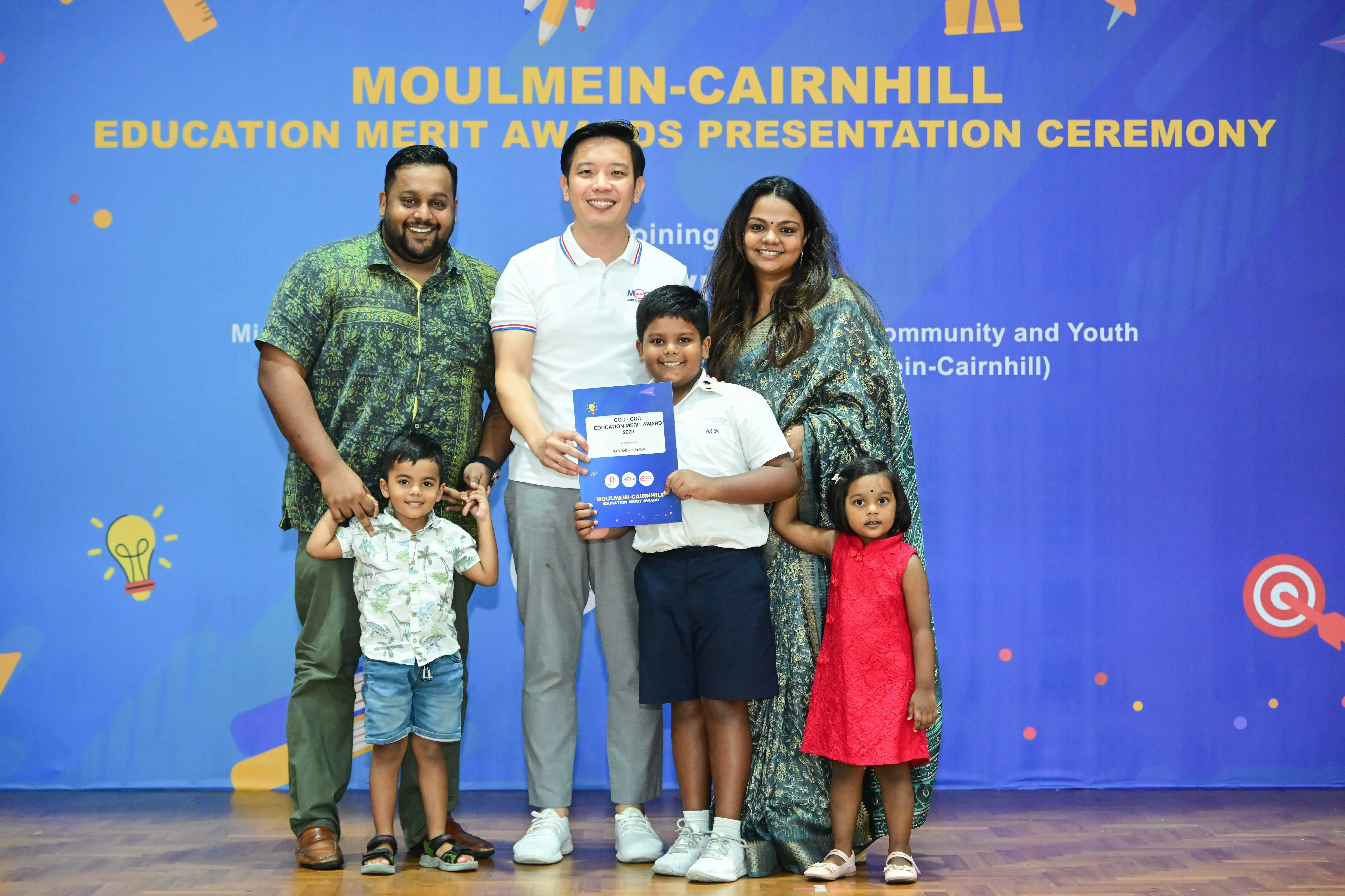 A family and an official pose with a merit award certificate at the Moulmein-Cairnhill Education Merit Awards ceremony.